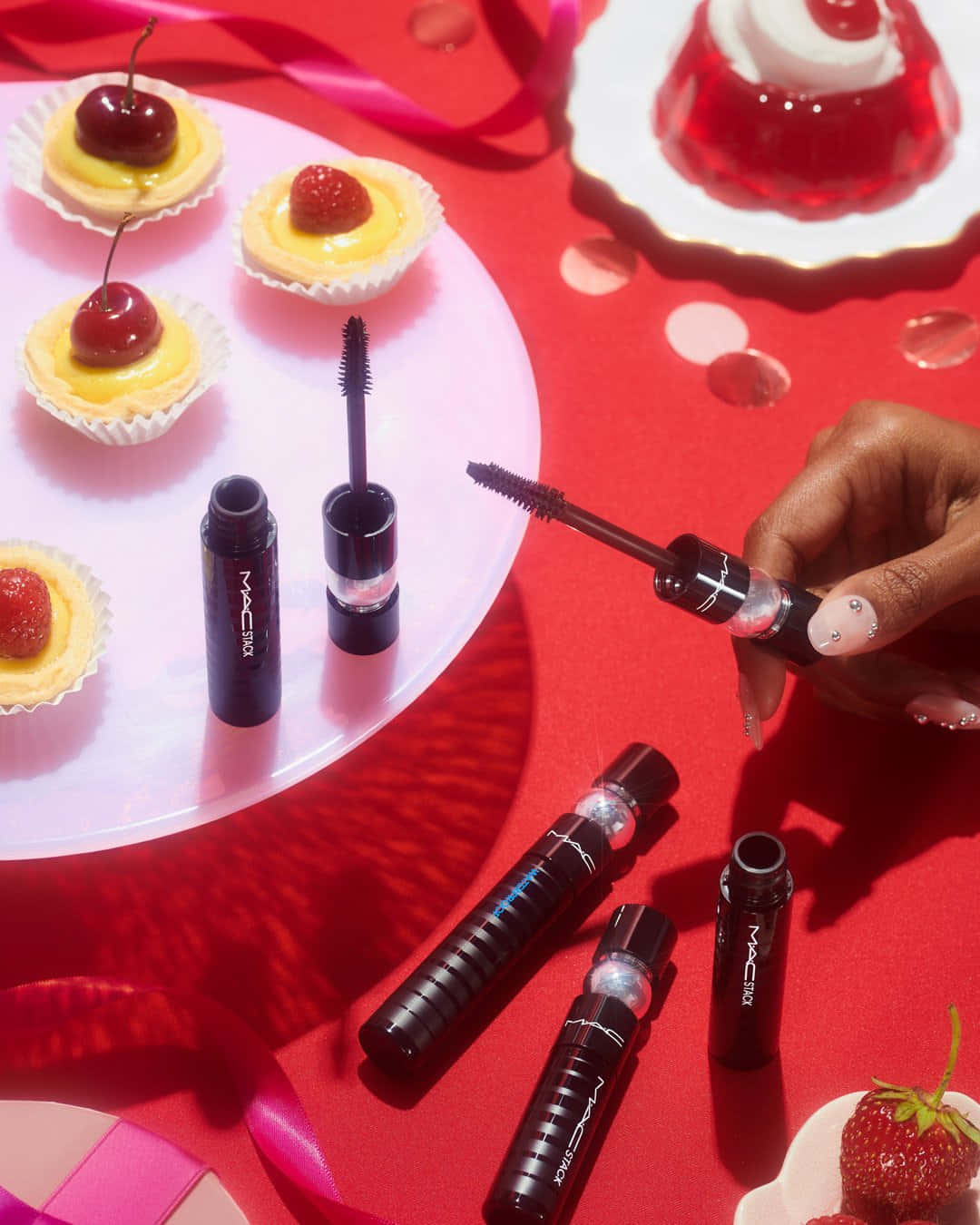 Mac Cosmetics 桌布 壁紙 — 一隻帶有閃亮指甲的手在一個散落著迷你塔、櫻桃和果凍甜點的鮮紅色表面上拿著一支MAC睫毛膏棒。
