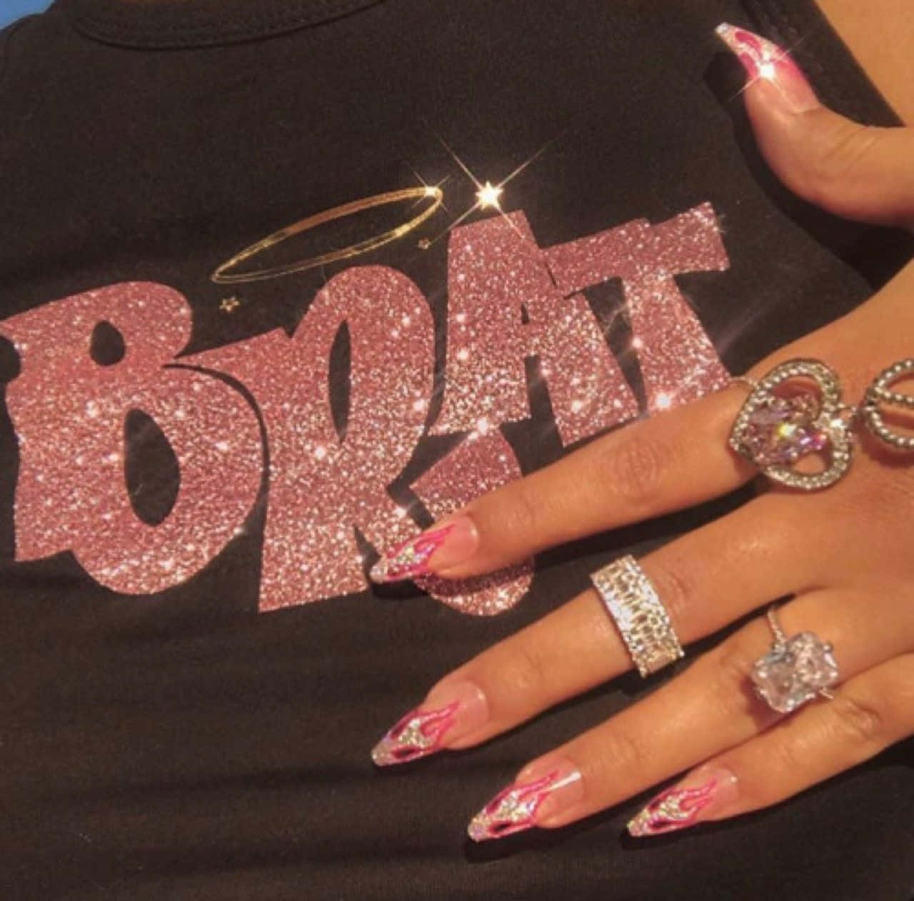 Pink Nails 桌布 壁紙 — 一隻帶有閃粉粉紅火焰指甲和鑽石戒指的手，放在一件帶有閃亮粉紅色「BRAT」標誌和光環的黑色襯衫上。