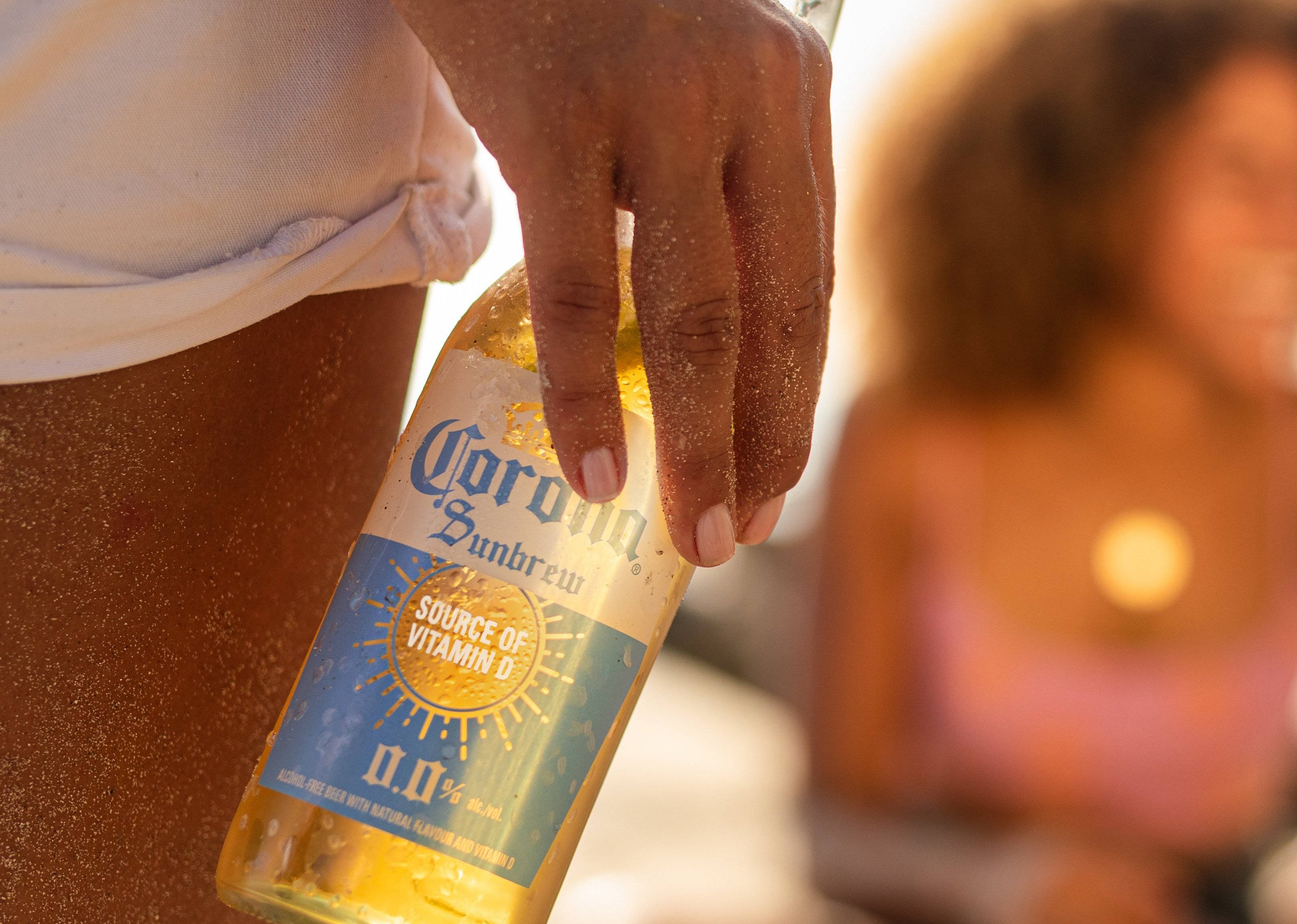 corona 桌布 壁紙 — 一隻帶沙的手拿著Corona Sunbrew瓶子，背景是陽光明媚的海灘，背景中有一個模糊的人影。