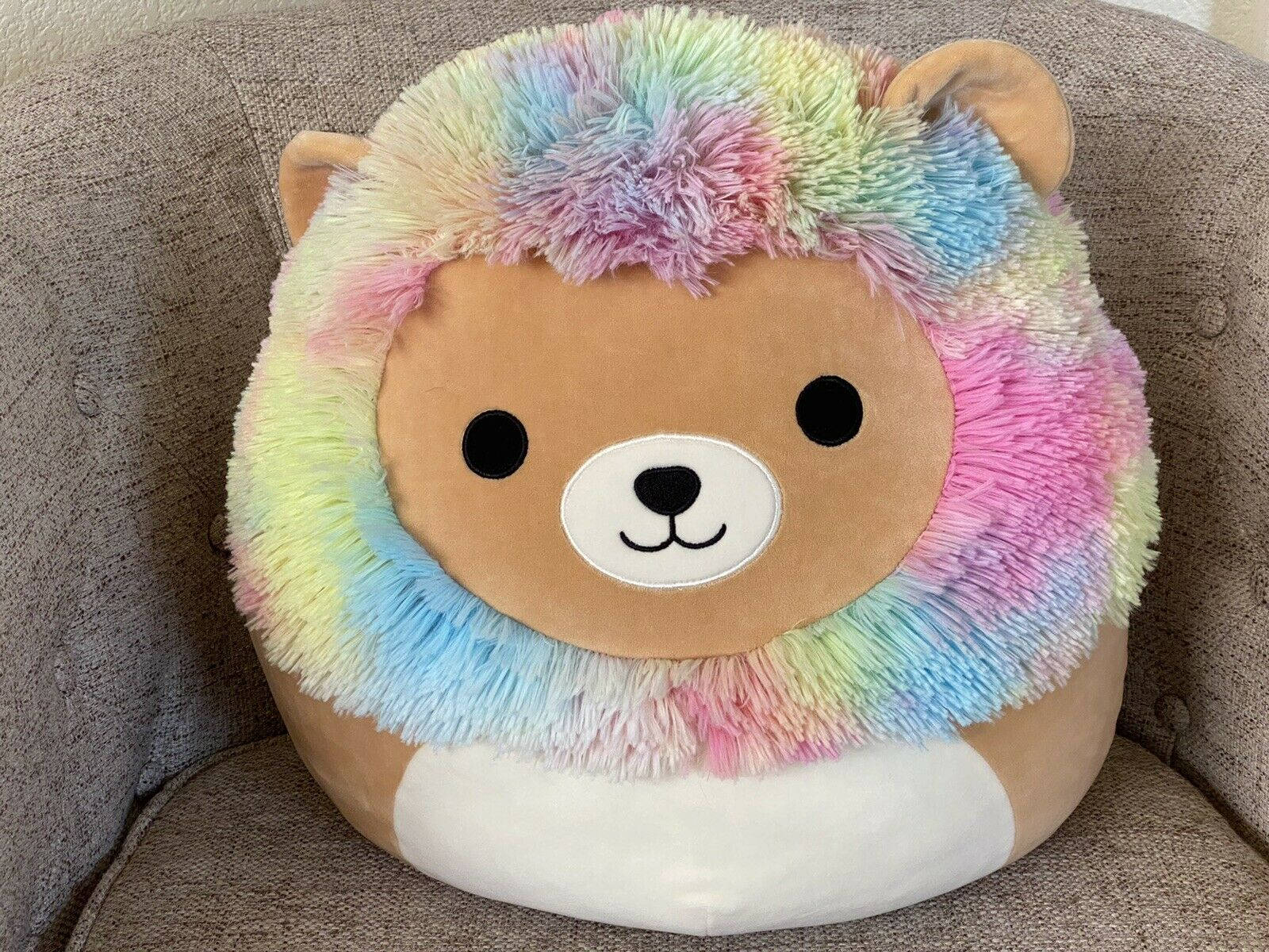 一隻彩虹毛絨獅子幼崽在灰色沙發上。 — Squishmallows 壁紙