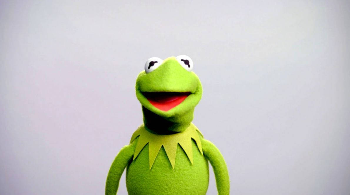 kermit 桌布 壁紙 — 一隻快樂的綠色青蛙木偶，有著寬闊的紅色嘴巴和黃色領子，在純灰色背景上微笑。