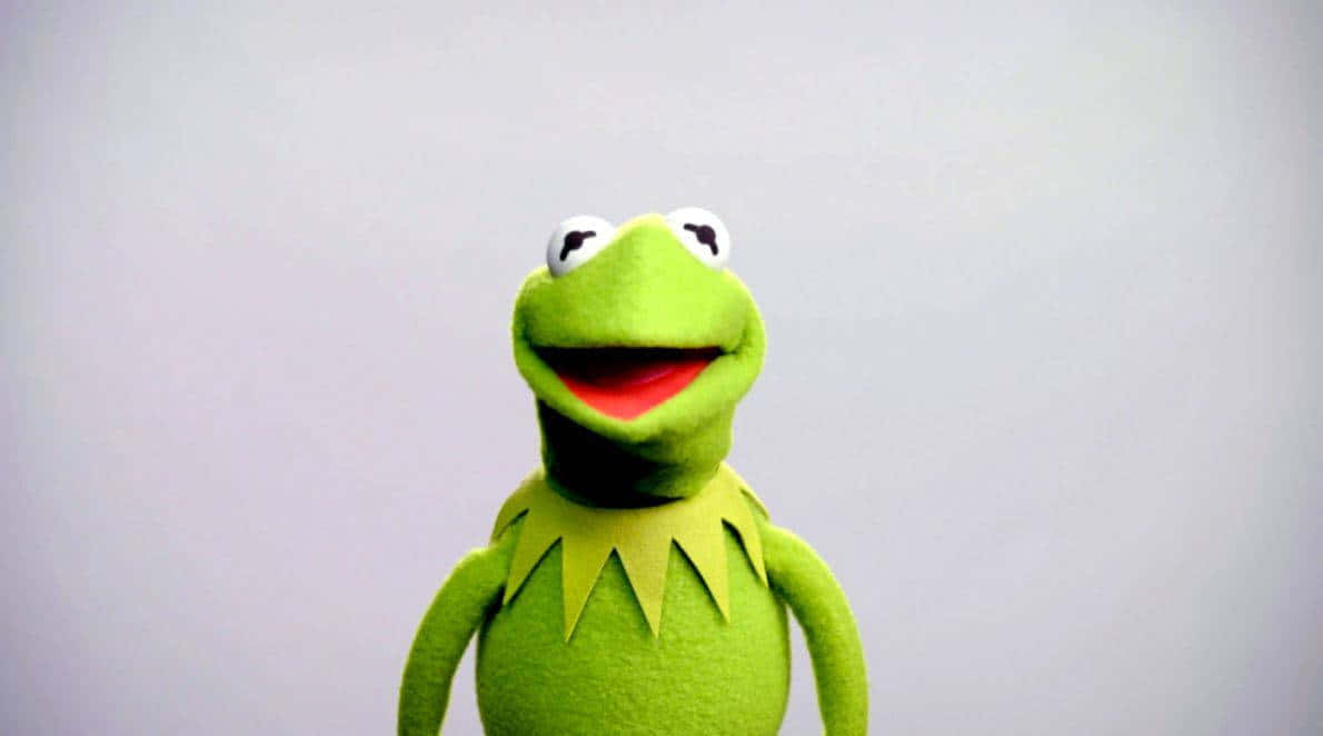 Aesthetic Kermit 桌布 壁紙 — 一隻快樂的綠色青蛙木偶，有著寬闊的紅色嘴巴和黃色領子，在純灰色背景上微笑。
