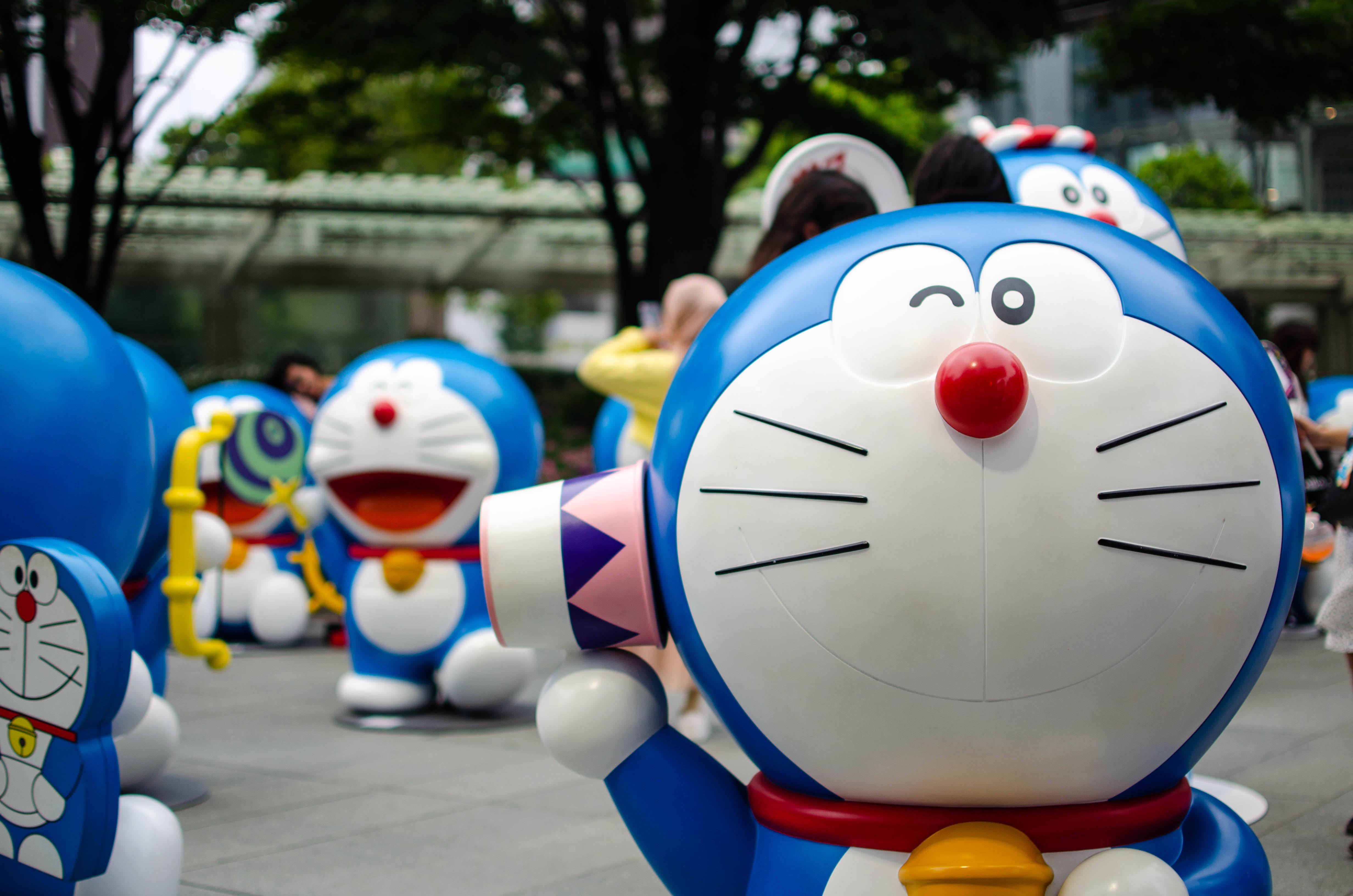 Doraemon 桌布 壁紙 — 一隻快樂的藍色機器貓，紅鼻子白臉，在陽光明媚的戶外廣場上，從一群相同的哆啦A夢雕像中揮手。