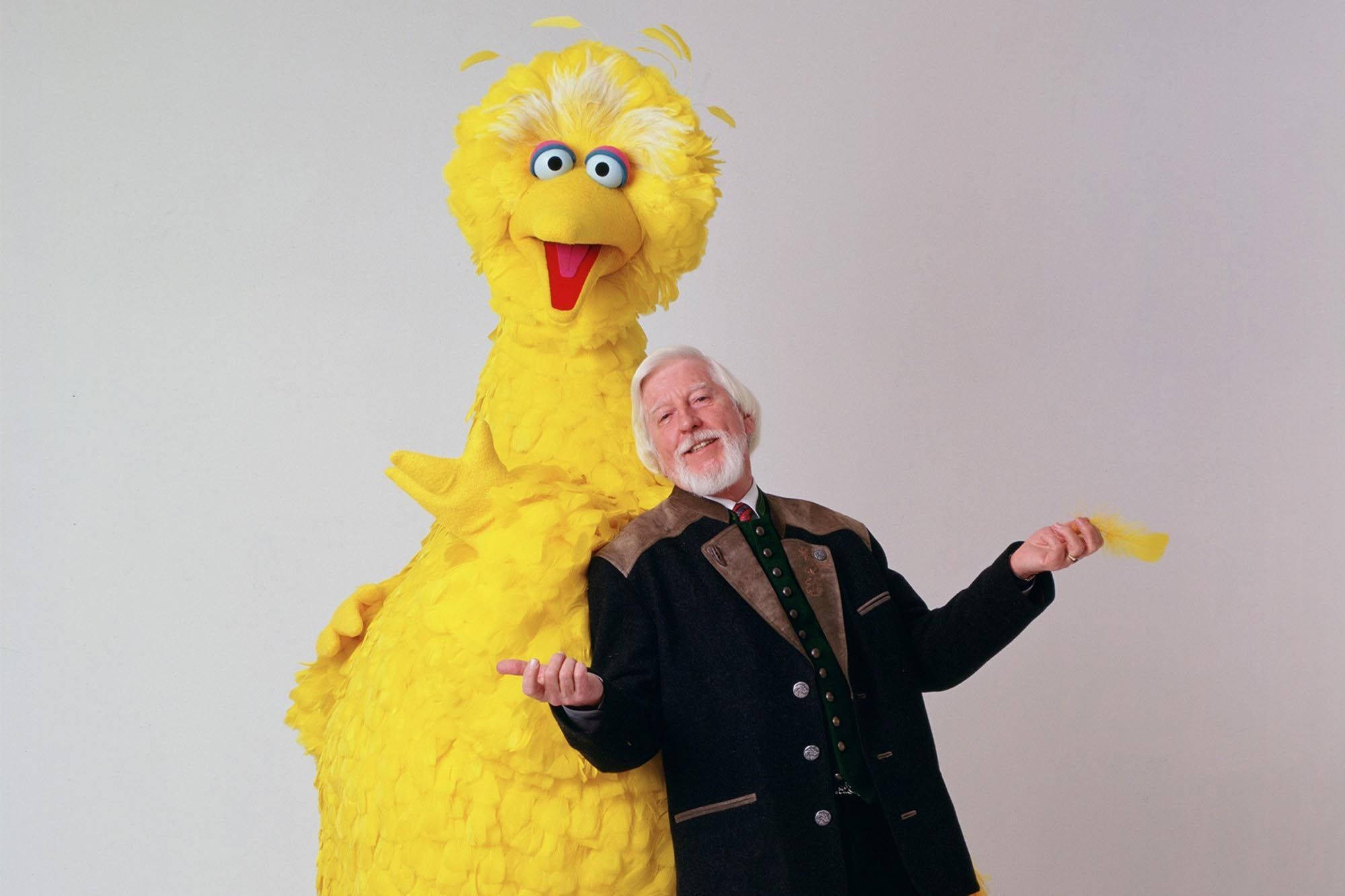 Big Bird 桌布 壁紙 — 一隻快樂的黃色大鳥布偶站在一位微笑的老年男子旁邊，男子身穿深色外套，背景簡單。