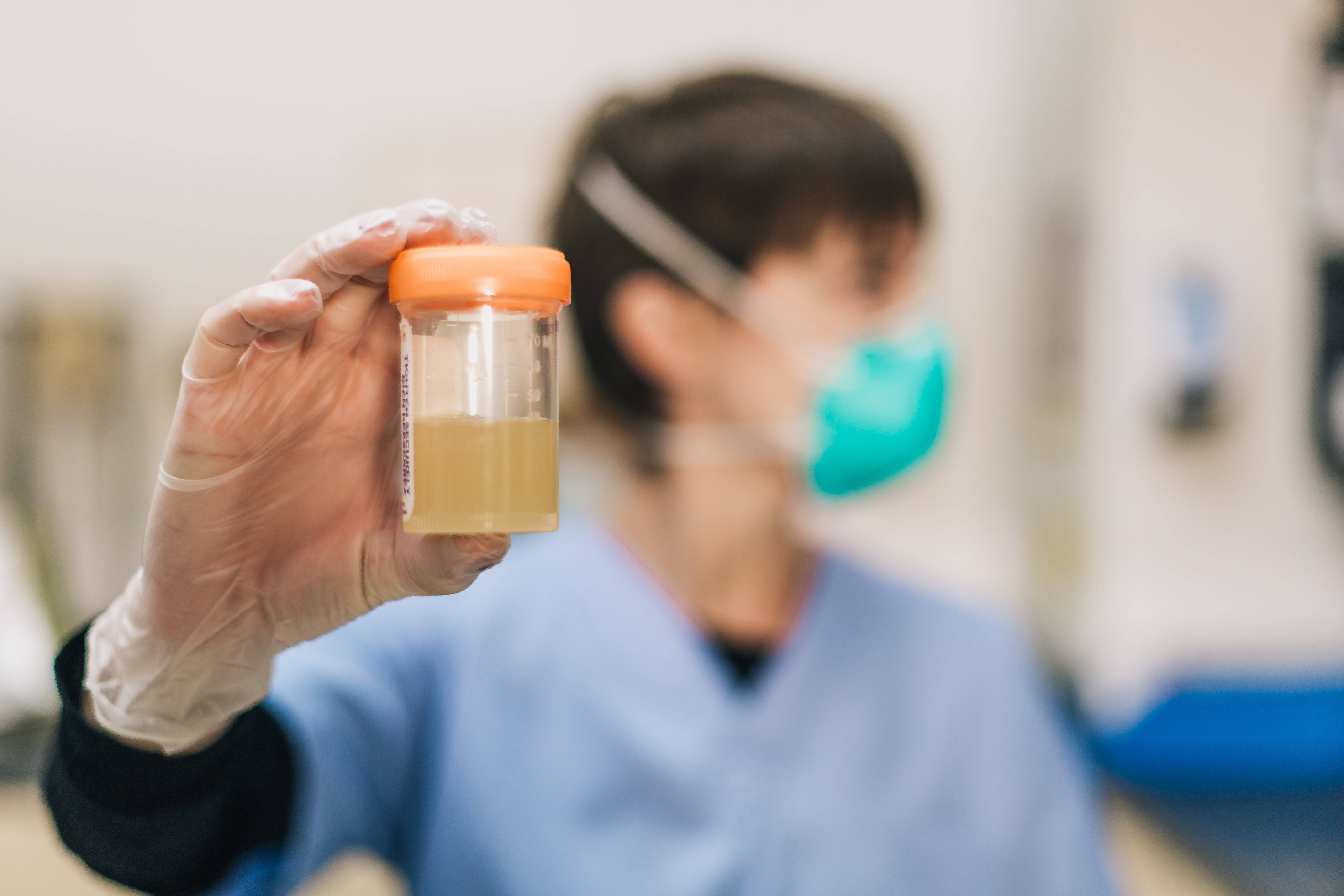 Medical 桌布 壁紙 — 一隻戴手套的手在臨床實驗室環境中拿著尿液樣本瓶，背景中有一位戴口罩的技術人員模糊不清。