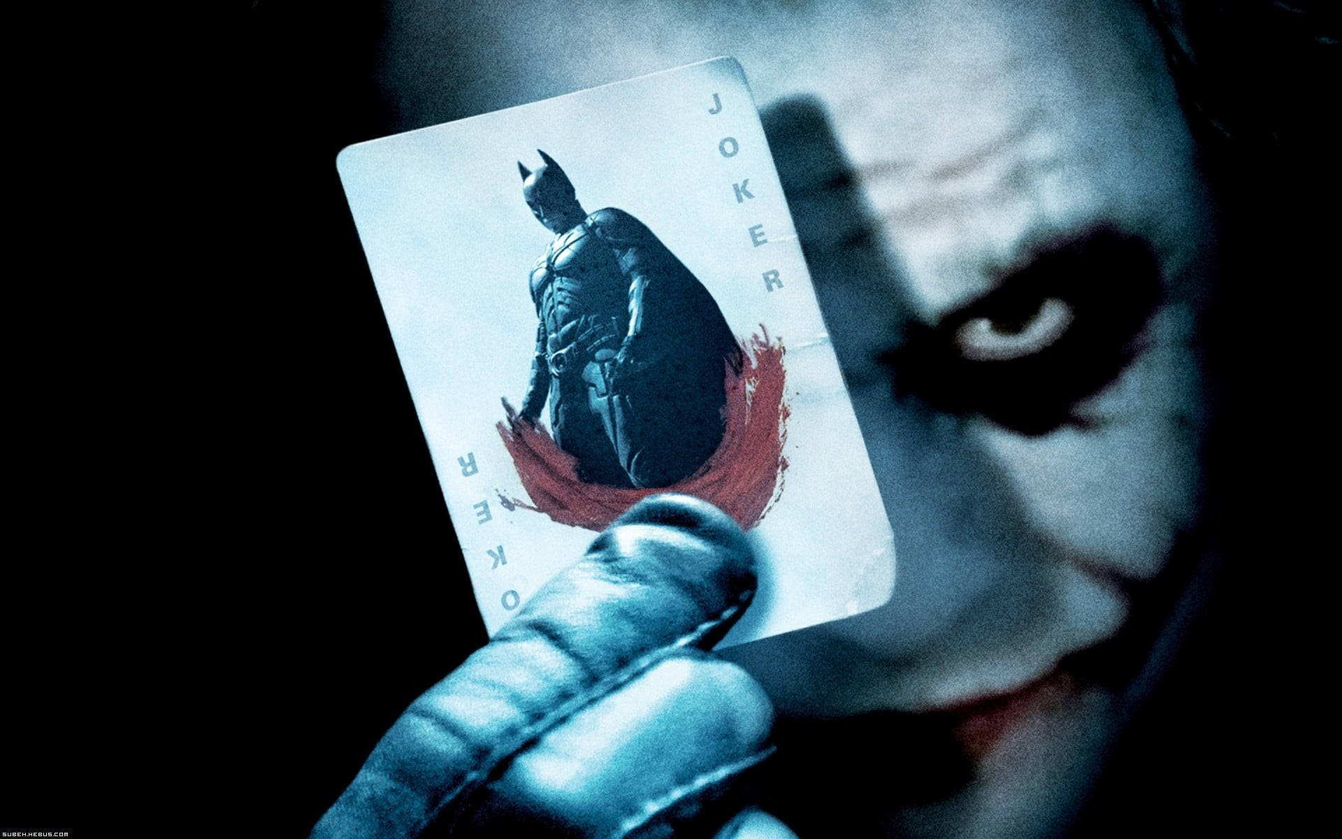Heath Ledger Joker 桌布 壁紙 — 一隻戴手套的手拿著一張印有蝙蝠俠的小丑撲克牌，背景是黑暗的，有一個帶威脅性的面孔部分可見。