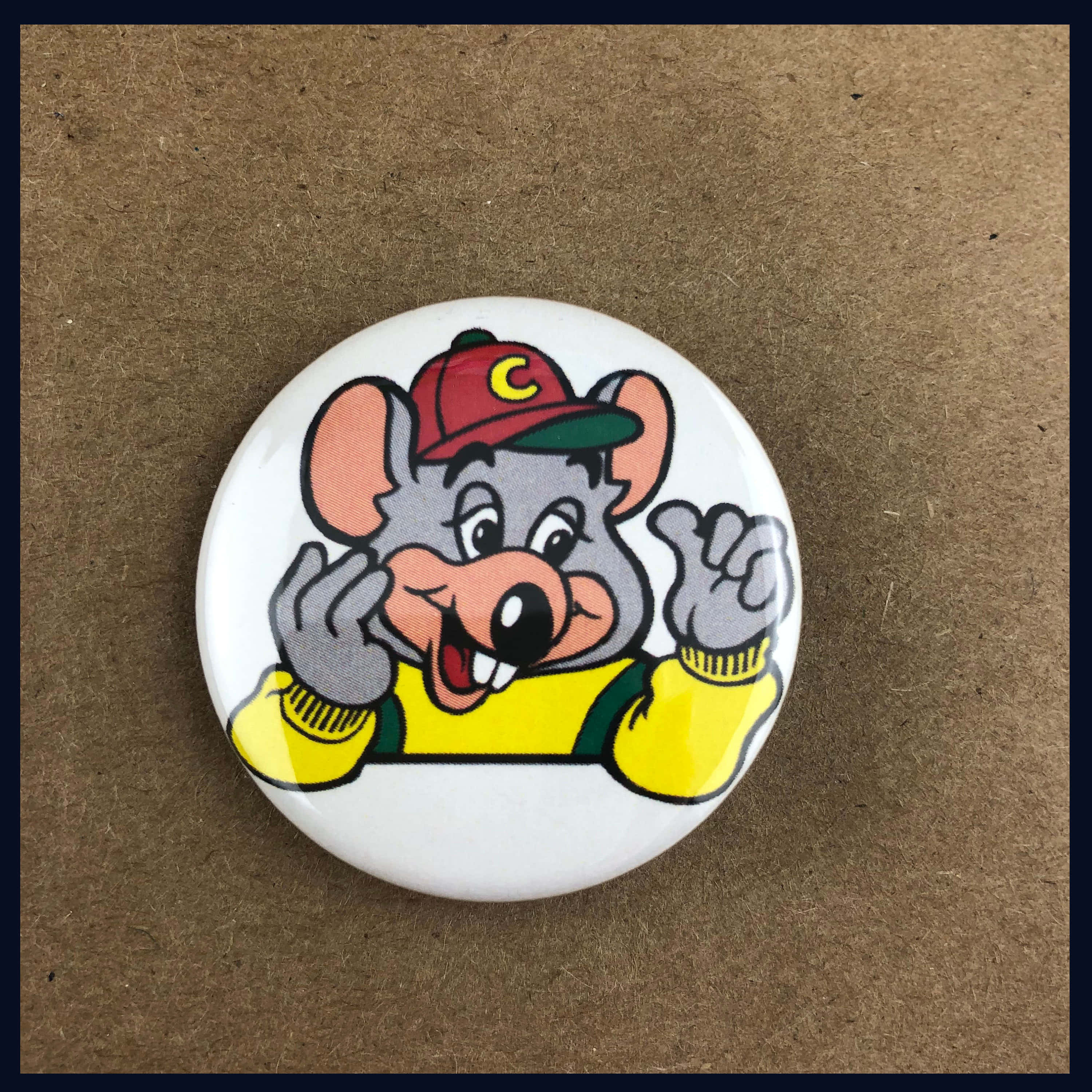 Chuck E Cheese 桌布 壁紙 — 一隻戴紅帽穿黃毛衣的開心卡通老鼠，在棕色紙板上的一個圓形白色鈕扣上微笑揮手。