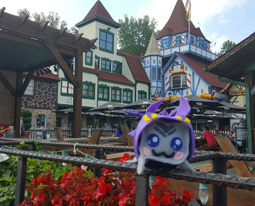 Vacation 桌布 壁紙 — 一隻戴紫色帽子的毛絨玩具在色彩繽紛的歐式建築前面，隔著欄杆偷看，建築物有紅色屋頂和戶外座位。