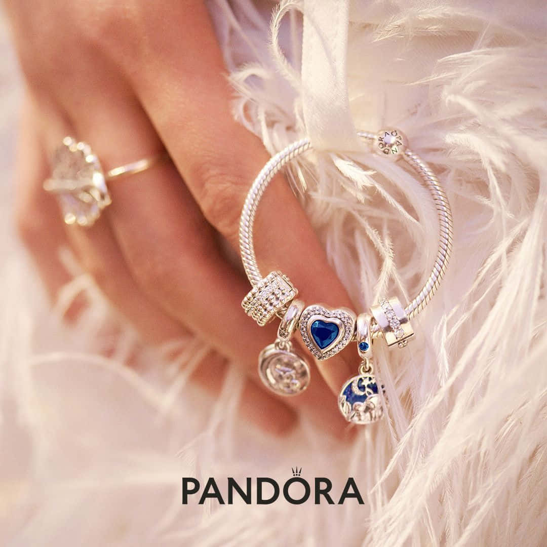 潘朵拉 桌布 壁紙 — 一隻戴著Pandora吊飾手鍊和戒指的手放在柔軟的白色羽毛上，展示閃爍的藍色和透明吊飾。
