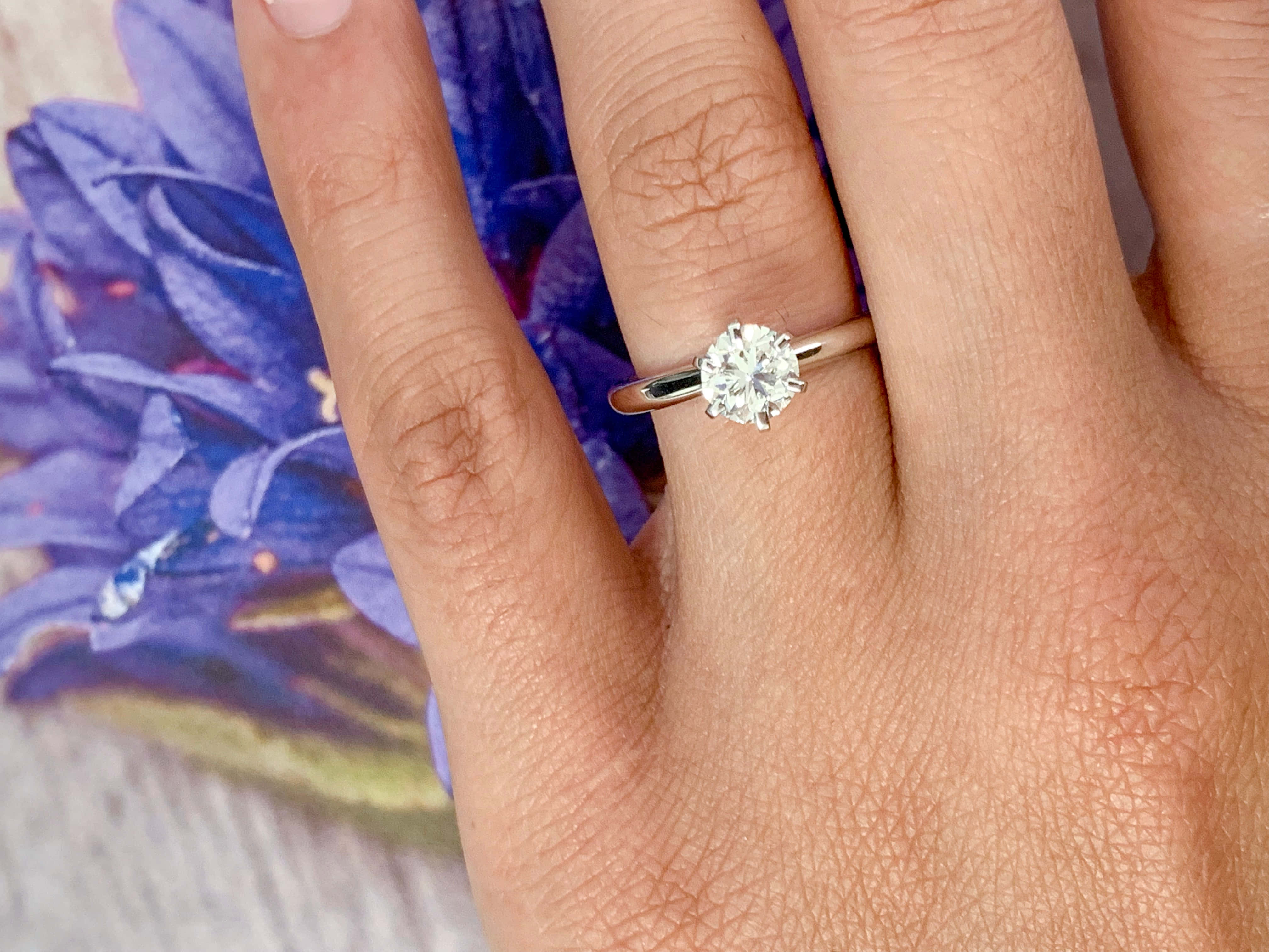 rings 桌布 壁紙 — 一隻戴著單鑽訂婚戒指的手，背景是模糊的紫色花卉。