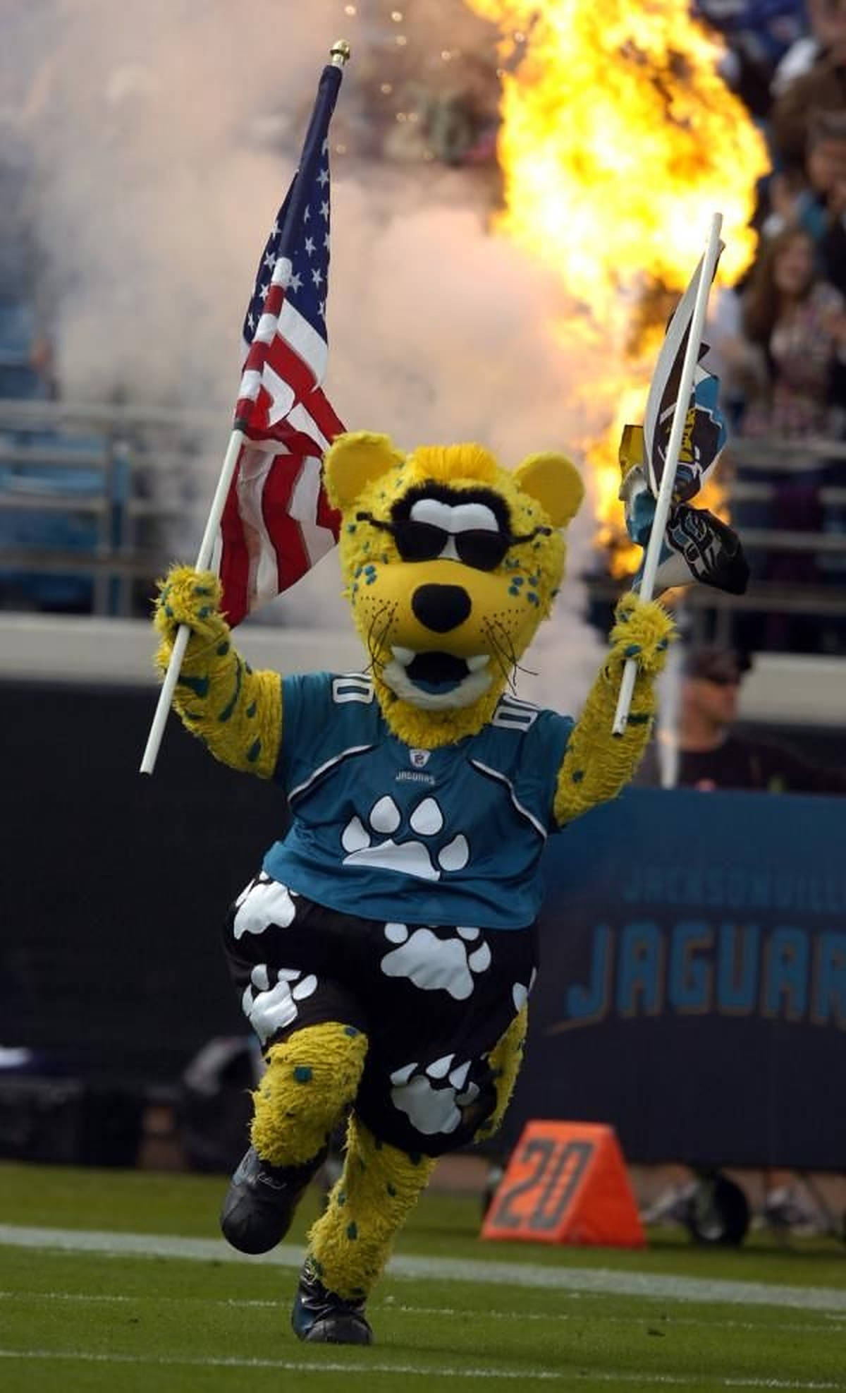 Jaguars 桌布 壁紙 — 一隻戴著太陽眼鏡和球隊裝備的黃色美洲虎吉祥物在足球場上精力充沛地奔跑，手持旗幟，背後有煙火爆炸。