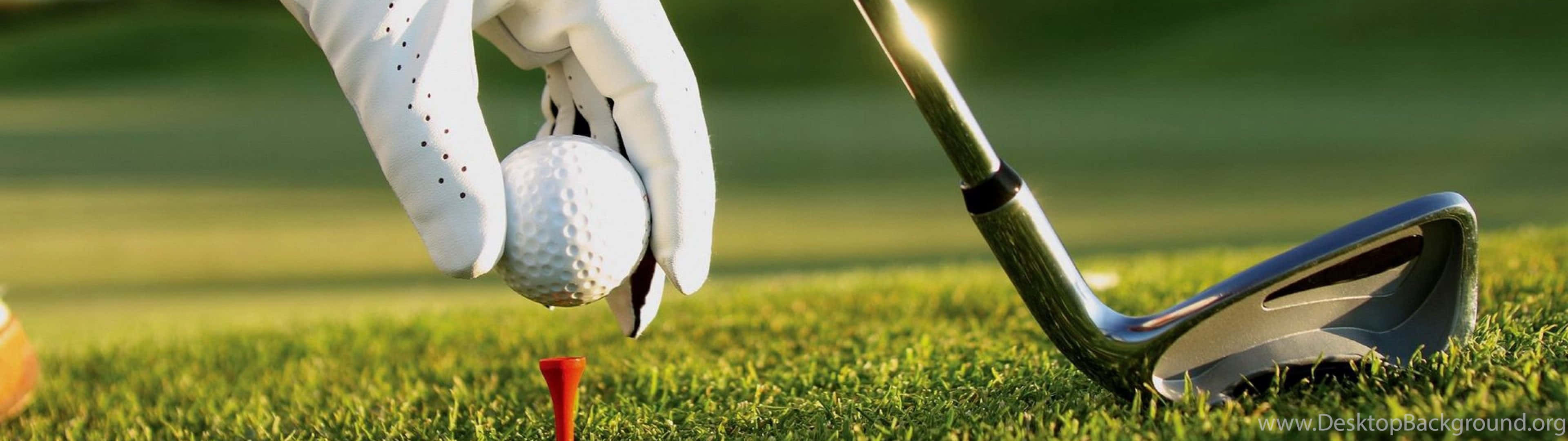 Golf 3840x1080 桌布 壁紙 — 一隻戴著手套的手，小心翼翼地將高爾夫球放在紅色球座上，旁邊是閃亮的球桿，背景是充滿活力的綠色草地。
