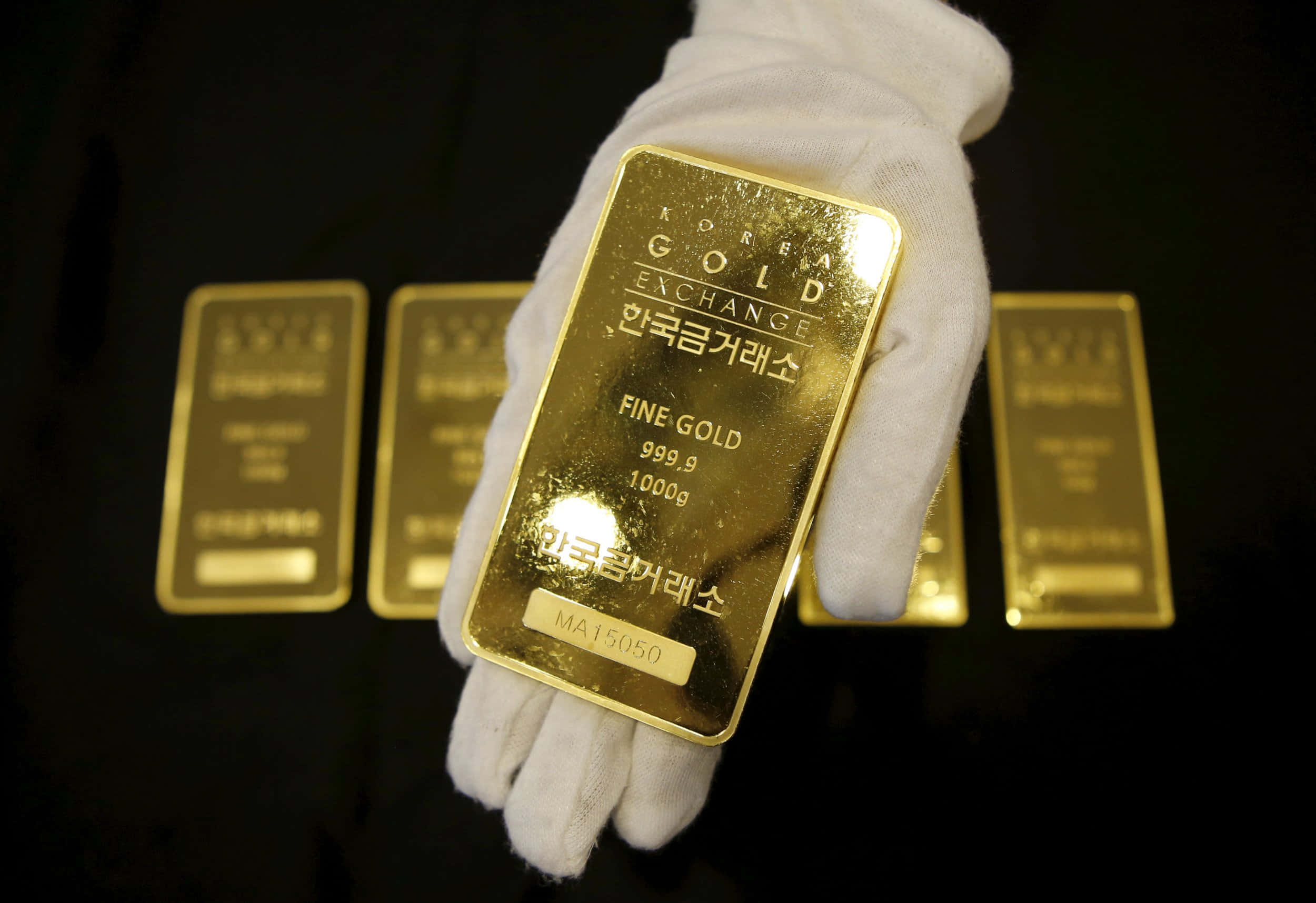 Gold Bar 桌布 壁紙 — 一隻戴著手套的手在深色背景上，與其他金條一起拿著一塊閃亮的1000g金條，上面標有Korea Gold Exchange。