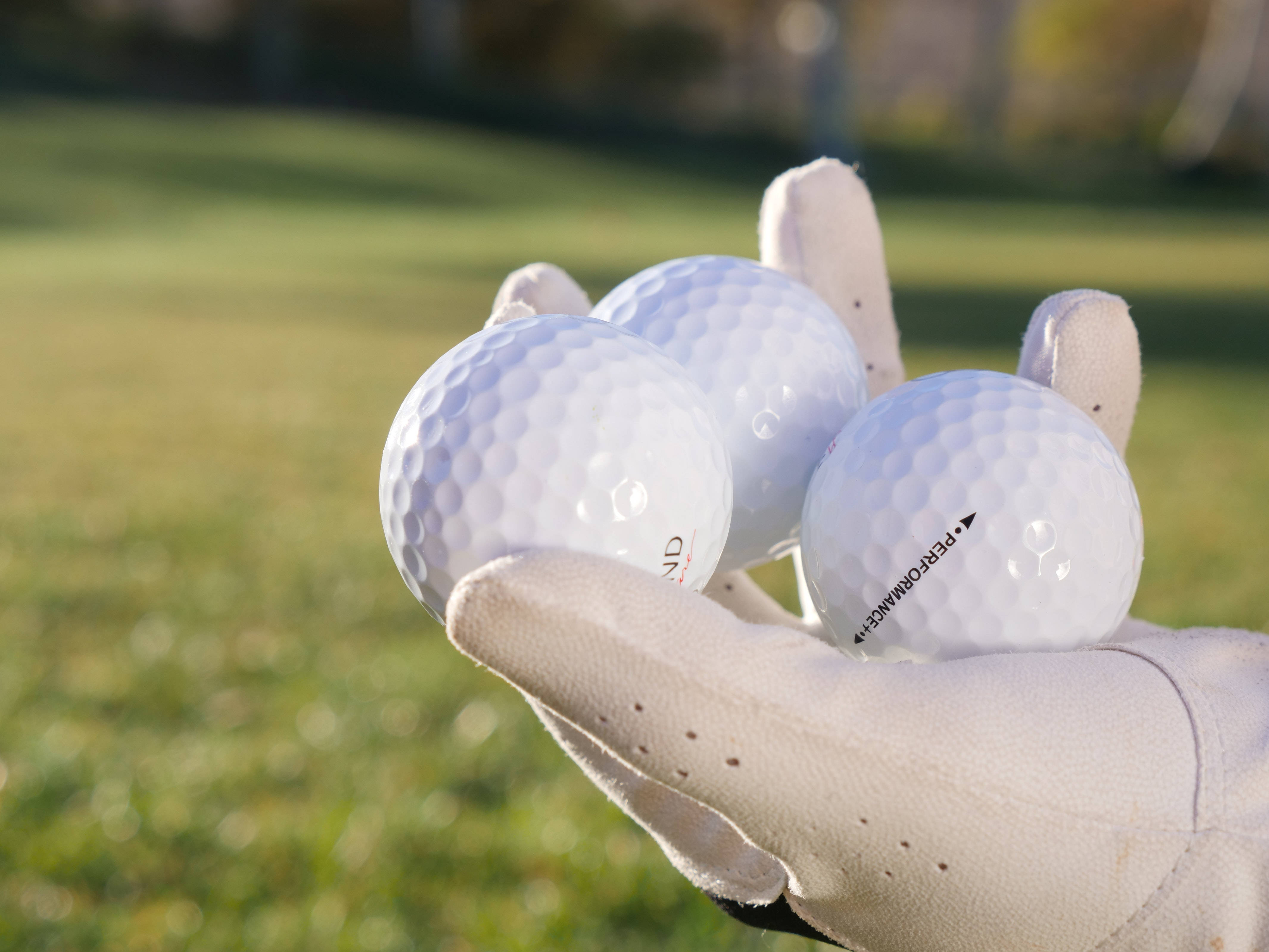 4K Golf 桌布 壁紙 — 一隻戴著手套的手在陽光普照的綠色草地背景下握著三個白色高爾夫球。
