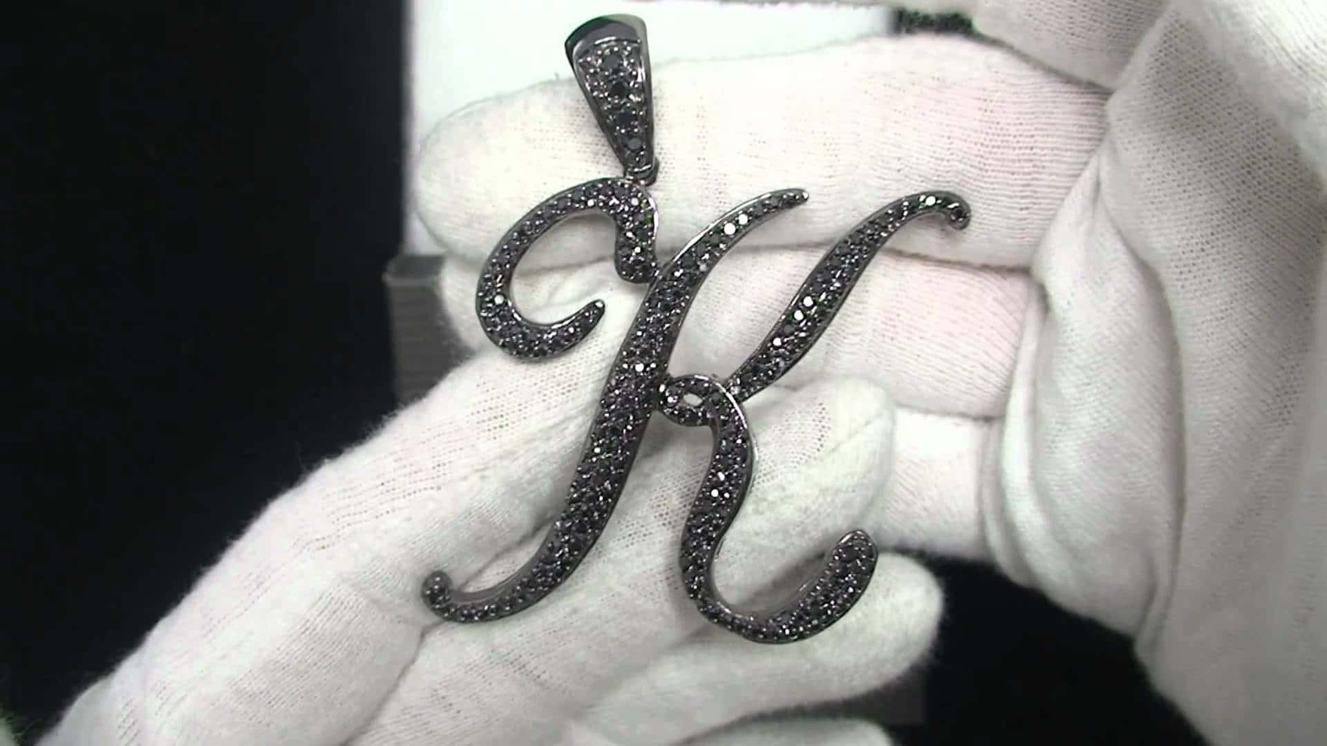 K 桌布 壁紙 — 一隻戴著白色手套的手，在深色背景前拿著一個閃閃發光、鑲有黑色鑽石的字母K吊墜。