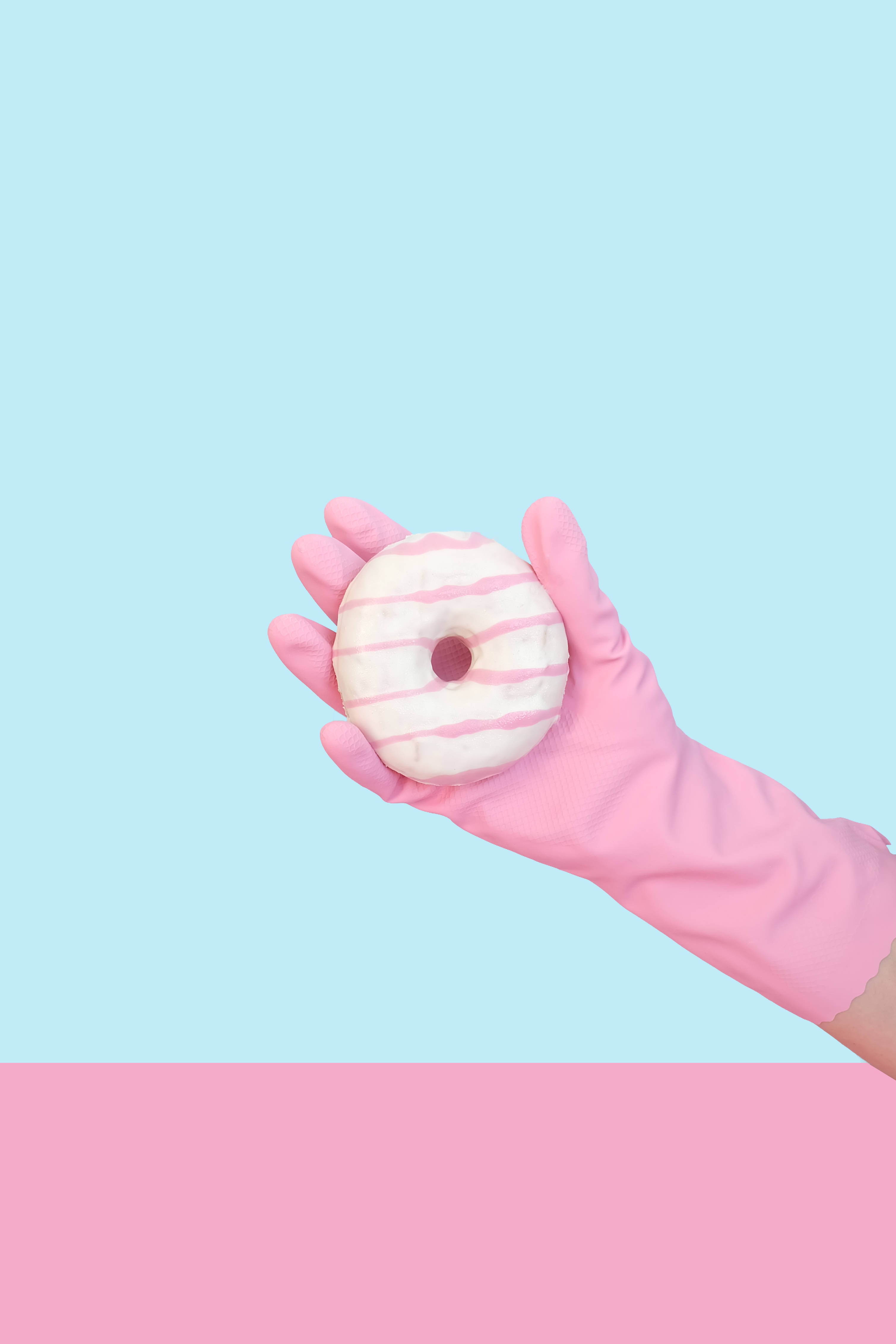Pastel Pink Aesthetic 桌布 壁紙 — 一隻戴著粉紅色橡膠手套的手，在粉藍色和粉紅色的粉彩背景前，拿著一個帶有粉紅色條紋的白色糖霜甜甜圈。