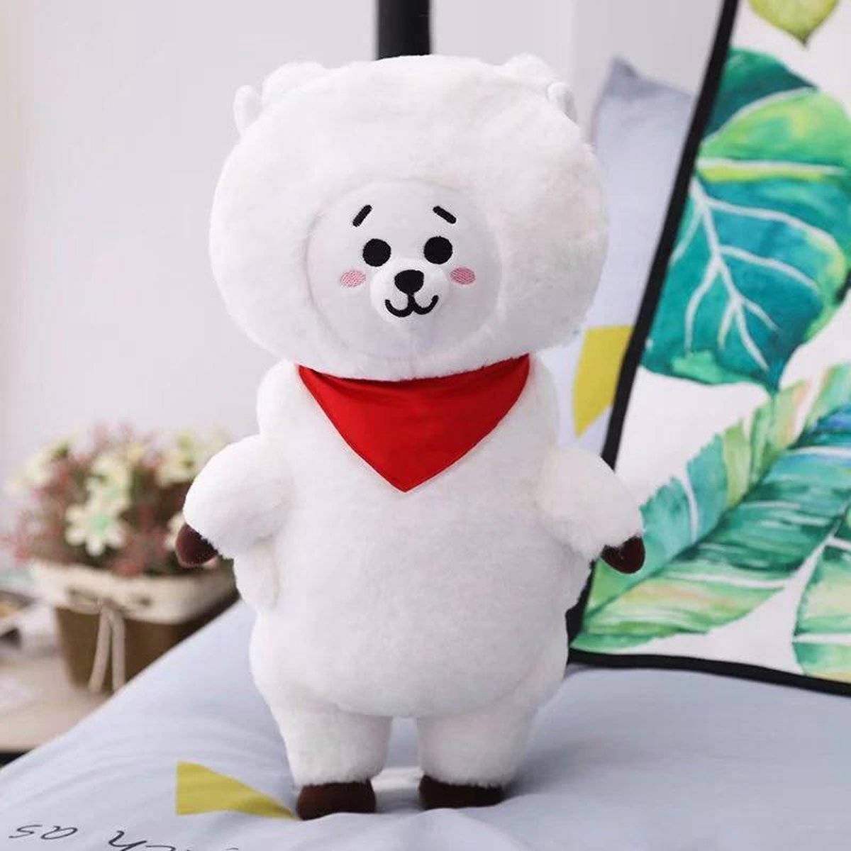 rj bt21 桌布 壁紙 — 一隻戴著紅色頭巾的 fluffy 白色毛絨熊站在床上，旁邊有一個熱帶葉子枕頭和一個盆栽植物。