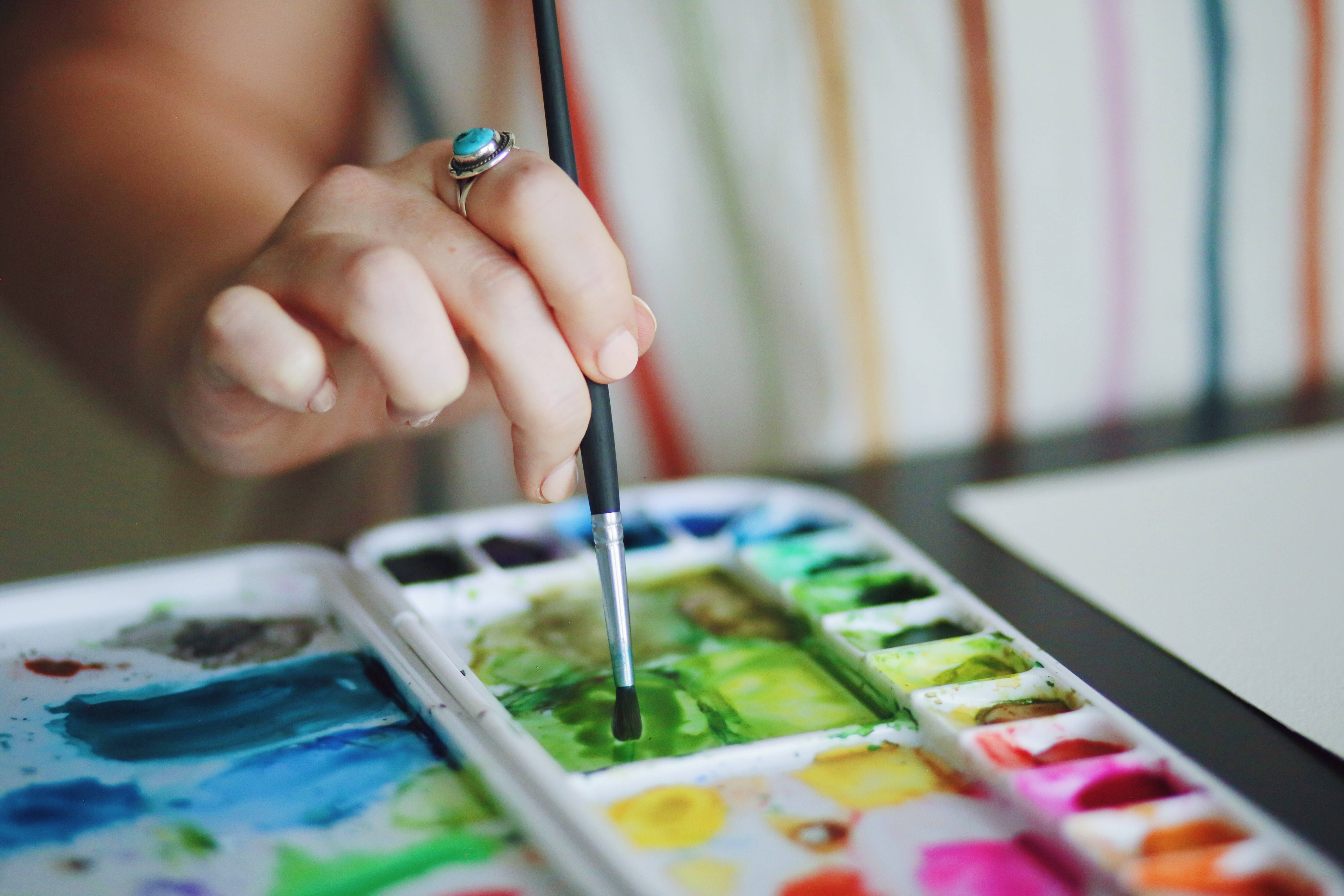 Watercolor 桌布 壁紙 — 一隻戴著綠松石戒指的手將畫筆浸入充滿鮮豔綠色、藍色和粉紅色調的水彩調色盤的特寫。