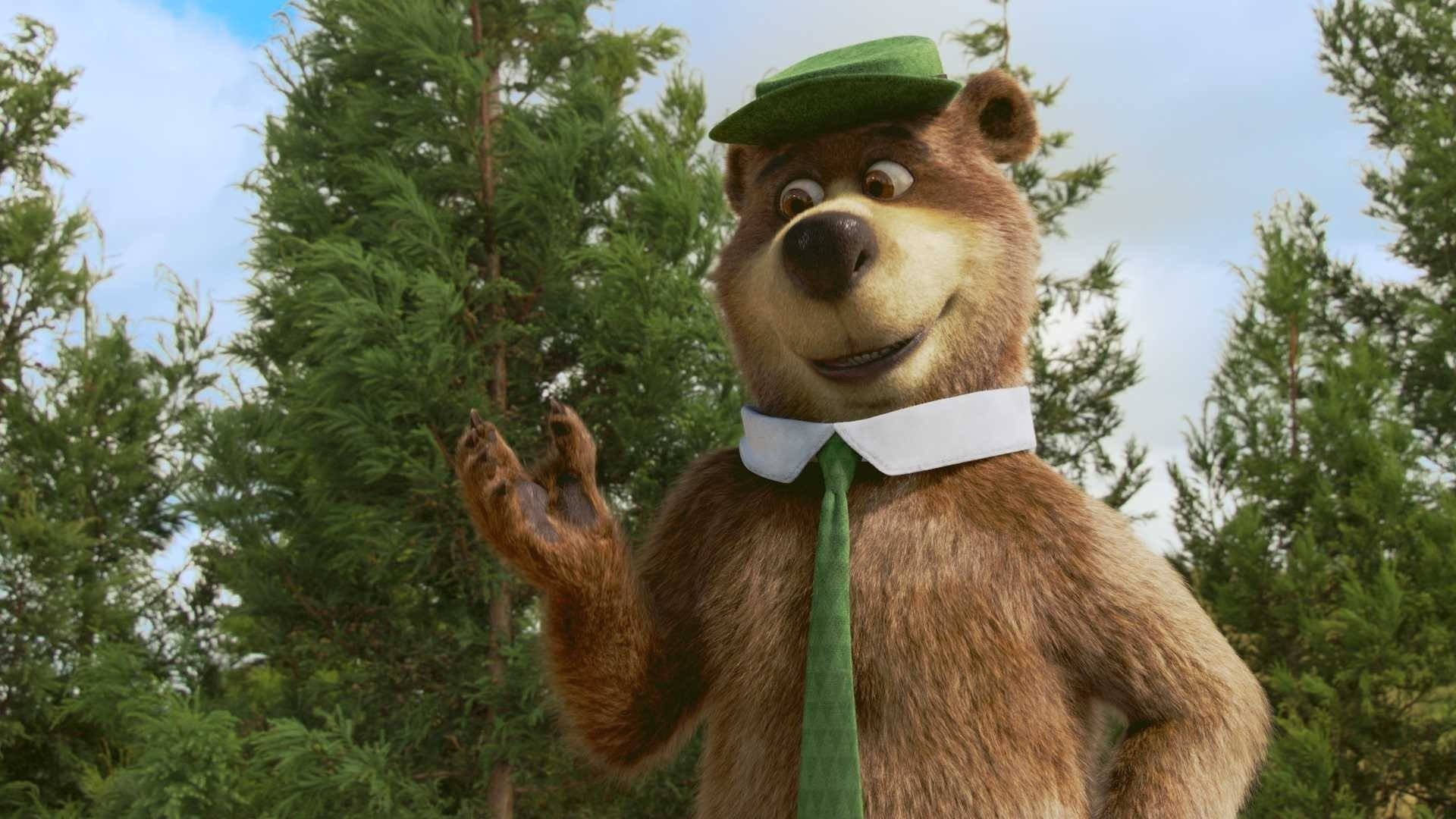Yogi 桌布 壁紙 — 一隻戴著綠色帽子和領帶的開心動畫熊在明亮的天空下，在高大的松樹間愉快地揮手。