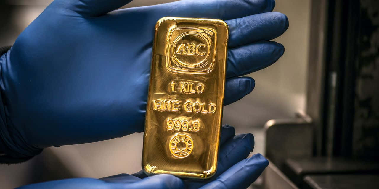 Gold Bars 桌布 壁紙 — 一隻戴著藍色手套的手在昏暗的工業環境中拿著一塊閃閃發光的1公斤純金金條，上面標有ABC 999.9。