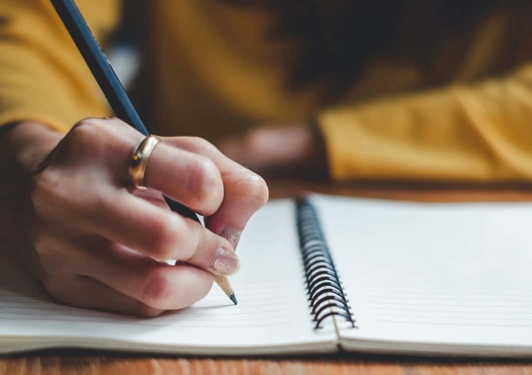 Writing 桌布 壁紙 — 一隻戴著金戒指的手在木桌上用藍色鉛筆在螺旋筆記本上寫字的特寫。