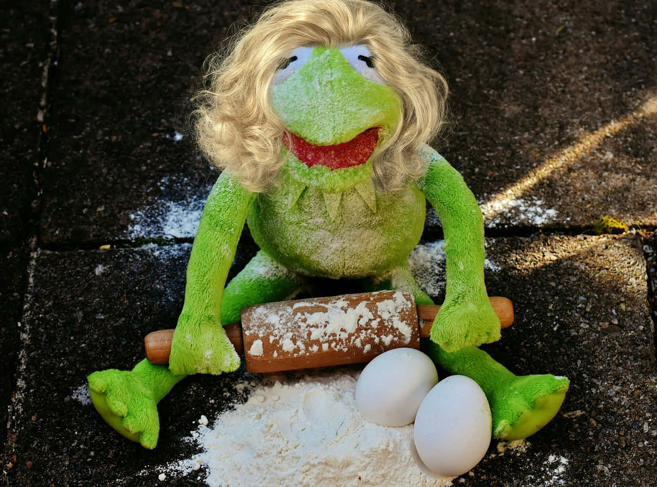 Eggs 桌布 壁紙 — 一隻戴著金色假髮的綠色青蛙玩偶，在石頭露臺上用擀麵棍揉麵粉，旁邊有兩個雞蛋。