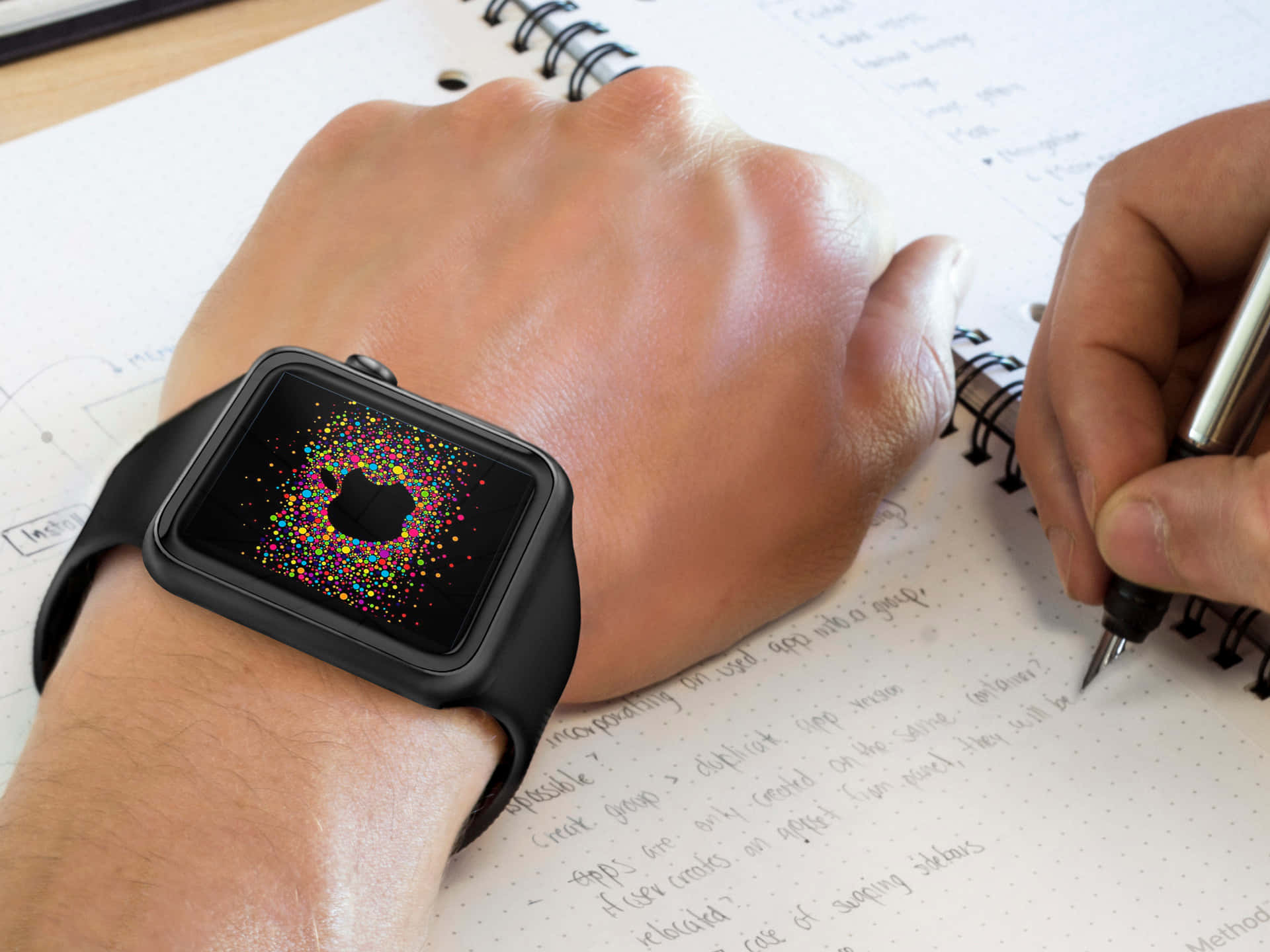 手錶 桌布 壁紙 — 一隻戴著黑色Apple Watch的手，螢幕上顯示色彩繽紛的粒子動畫，同時在筆記本上書寫的特寫。