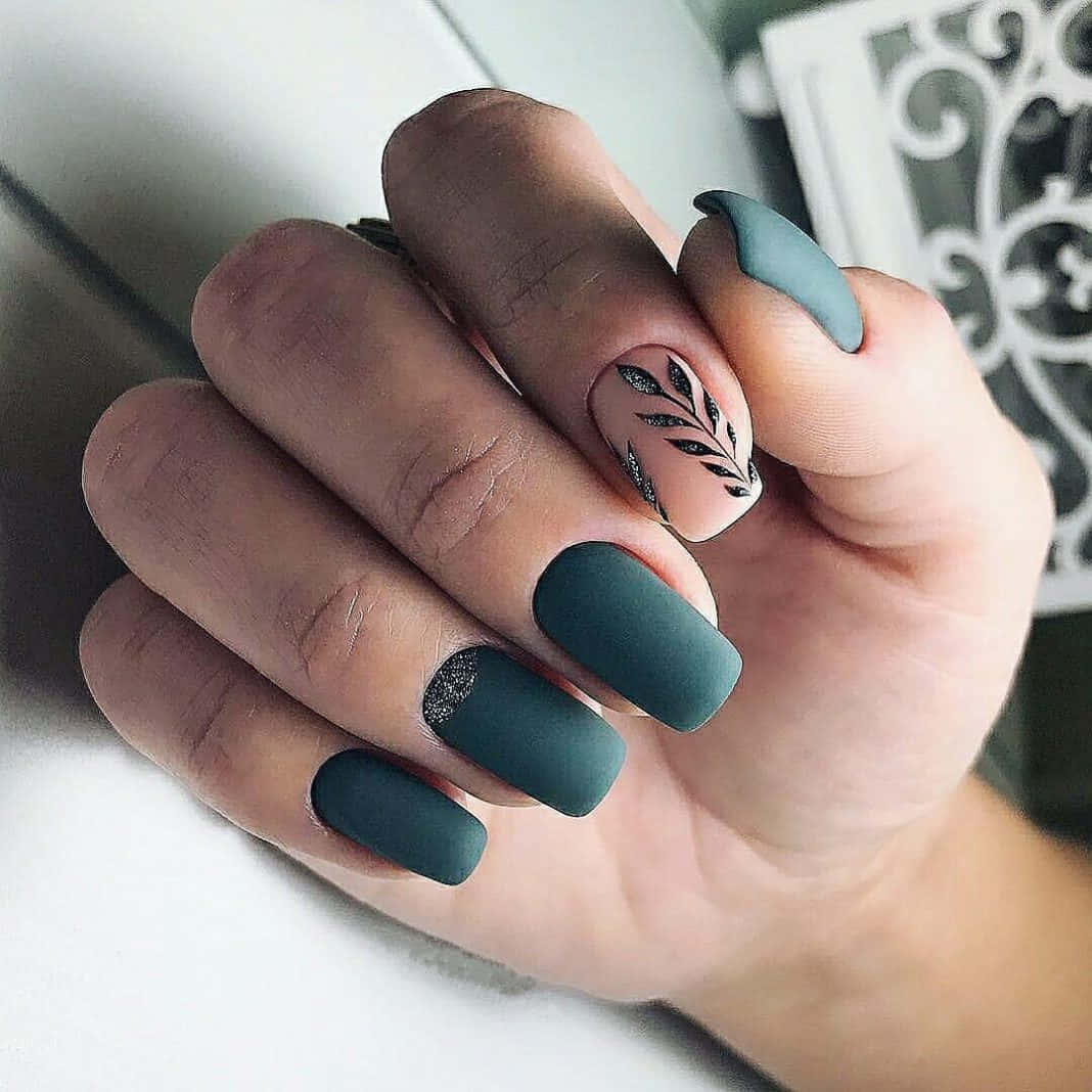 Spring Nails 桌布 壁紙 — 一隻手，帶有啞光青綠色指甲，飾有精緻的黑色葉子圖案和亮粉點綴，置於柔軟的白色表面上。