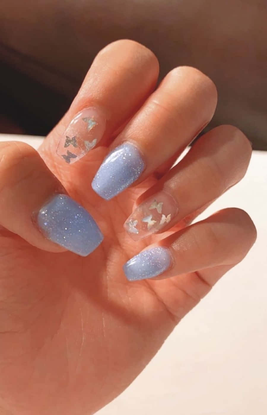 Nail 桌布 壁紙 — 一隻手，帶有淺藍色亮粉指甲和透明指尖，裝飾著細小的銀色蝴蝶。