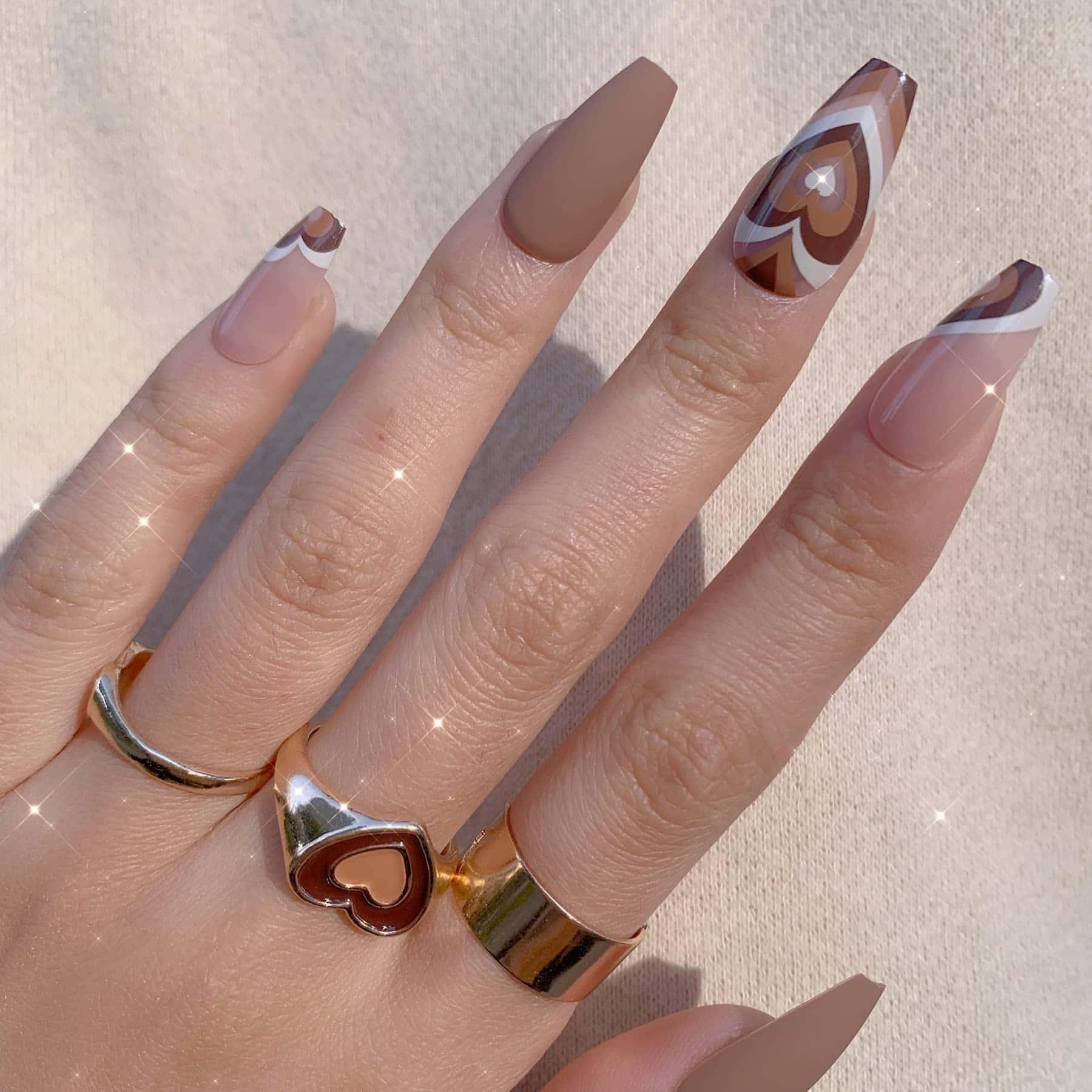 Fake Nail 桌布 壁紙 — 一隻手，長有杏仁形指甲，帶有棕色和白色的幾何心形圖案，金色戒指，背景為柔和的米色。