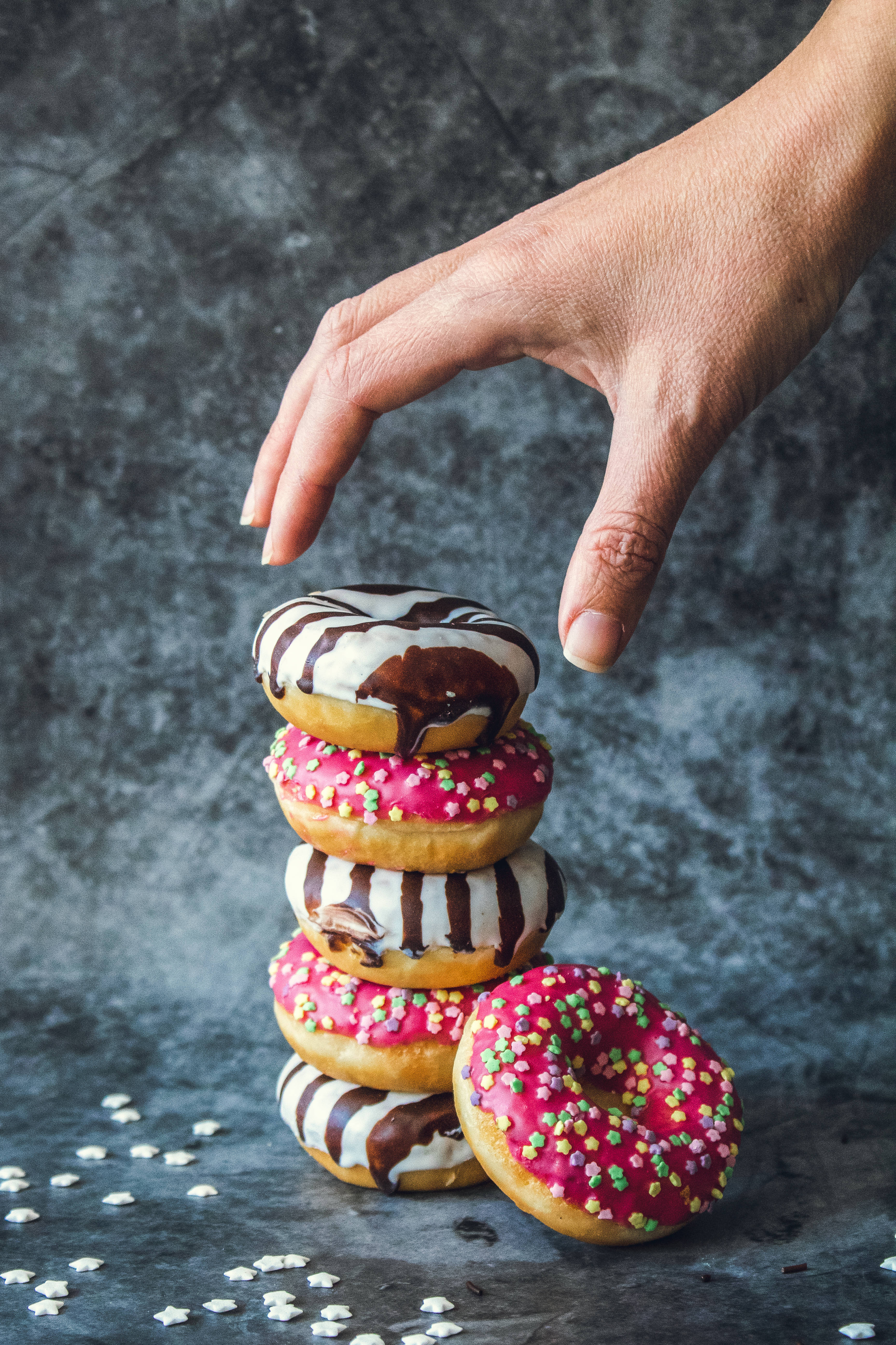 Dunkin Donuts 桌布 壁紙 — 一隻手伸向一堆色彩繽紛的糖霜甜甜圈，上面有粉紅色糖粒和巧克力醬，背景是深色紋理。