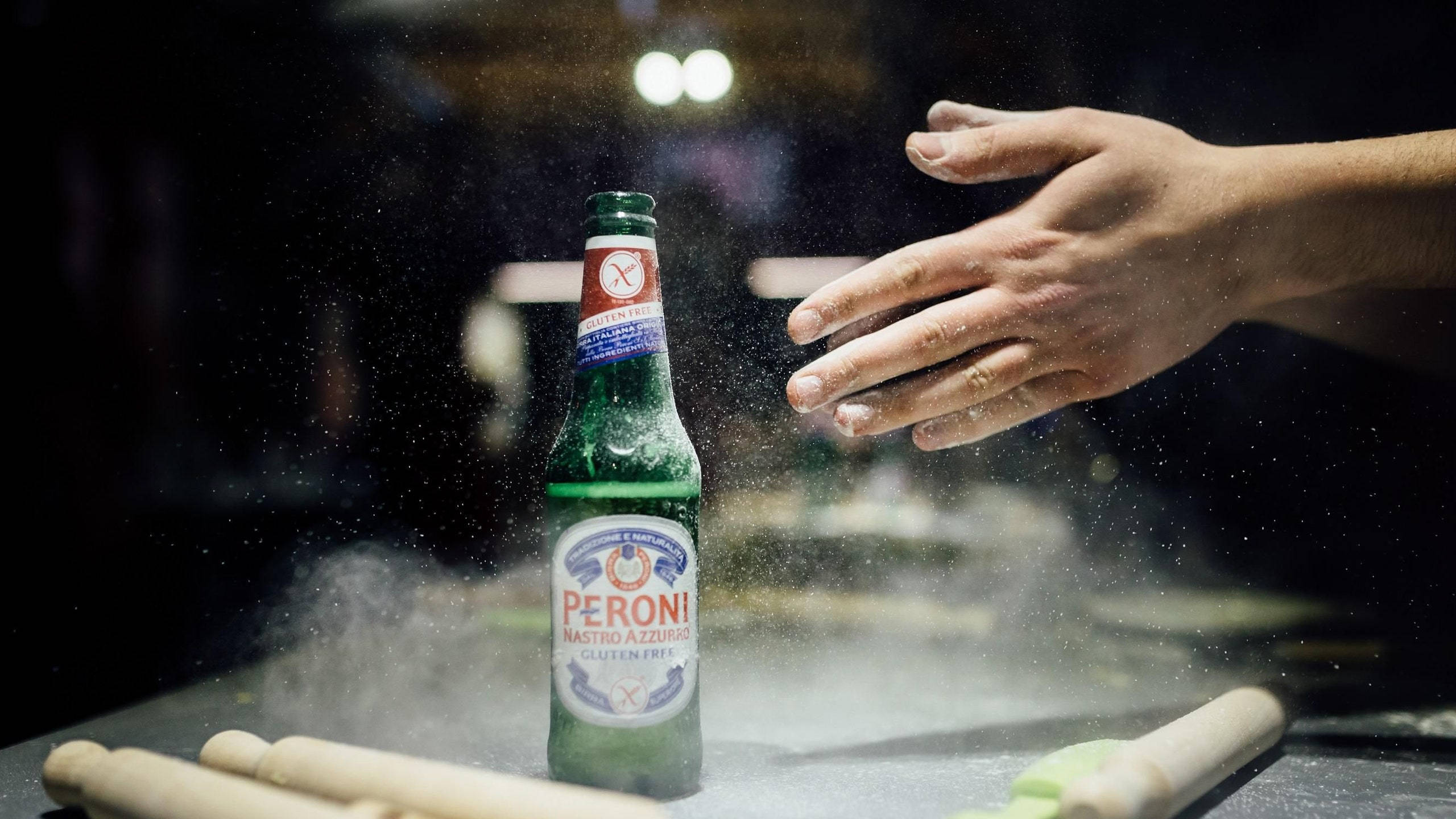 bottle 桌布 壁紙 — 一隻手伸向被麵粉和擀麵棍包圍的 Peroni Nastro Azzurro 無麩質啤酒瓶，廚房燈光昏暗。