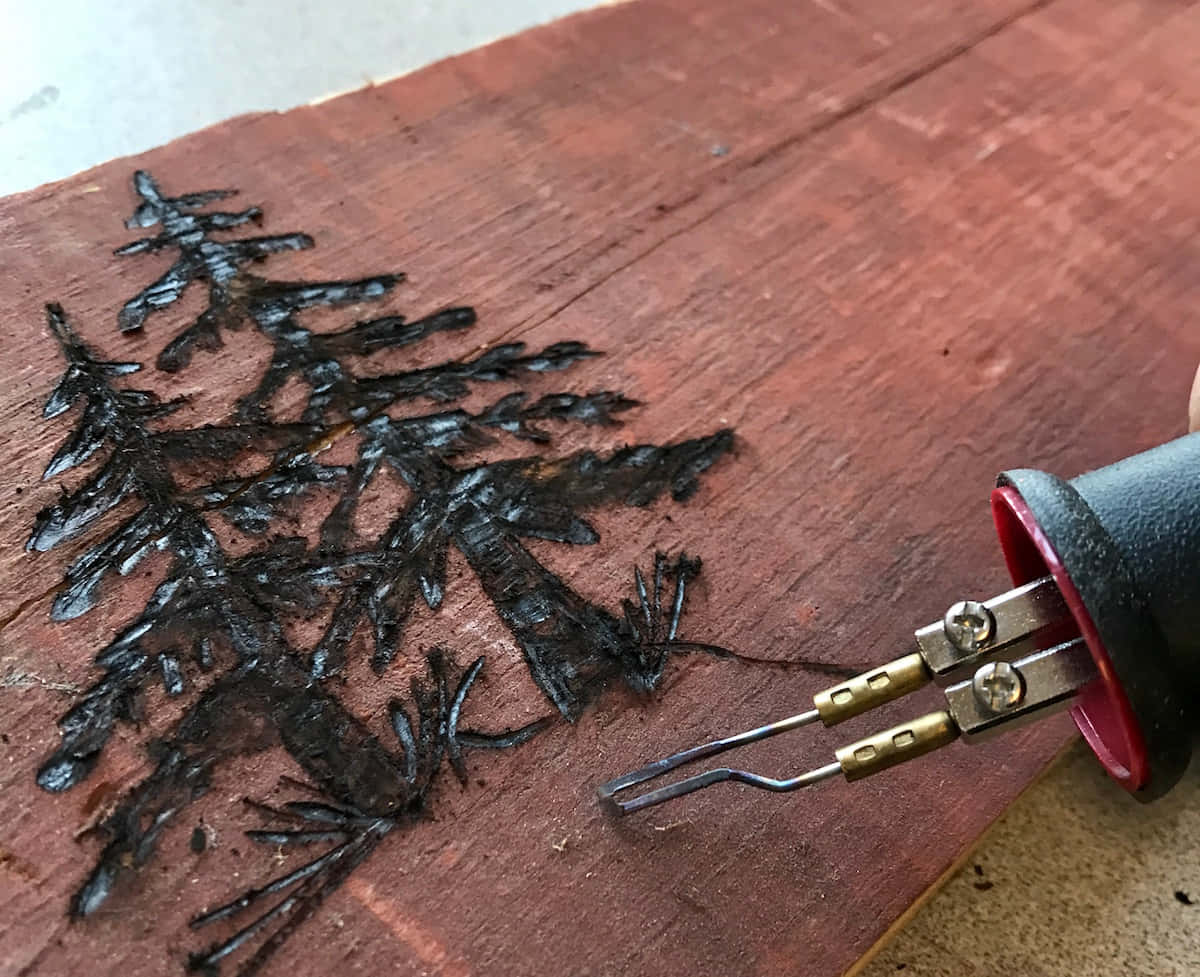 Wood Burning 桌布 壁紙 — 一隻手使用木燒工具在豐富的紅褐色木板上蝕刻出詳細的松樹圖案。