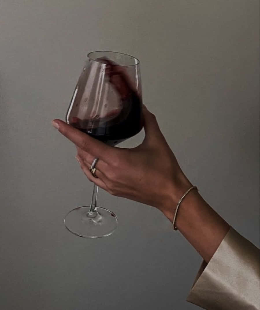 Black Wine 桌布 壁紙 — 一隻手優雅地握著一杯深紅葡萄酒，背景是柔和的灰色，光線柔和。
