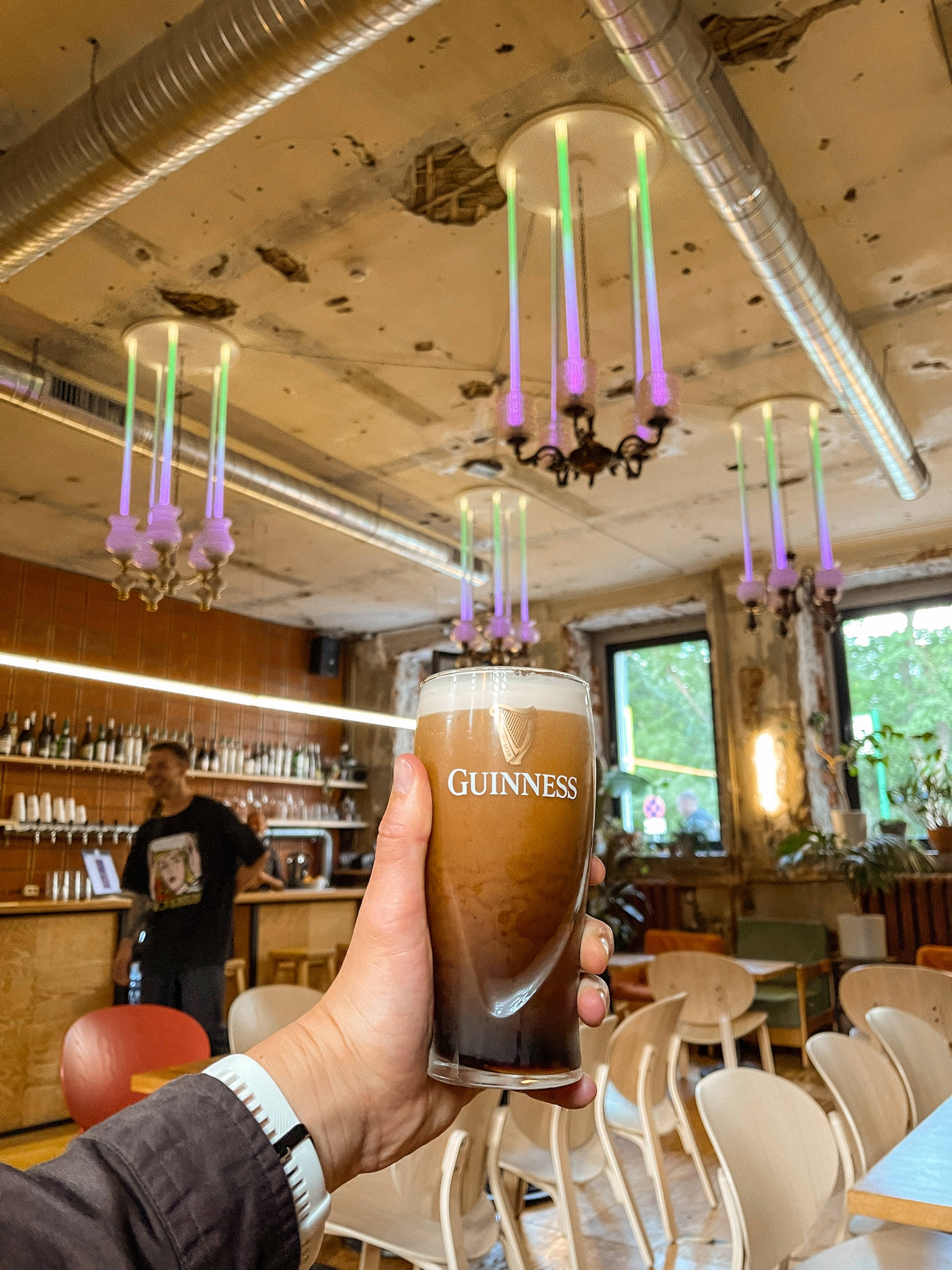 Beer 桌布 壁紙 — 一隻手在一個帶有彩色吊燈和裸露管道的鄉村工業風格酒吧中，拿著一杯泡沫豐富的健力士啤酒杯。