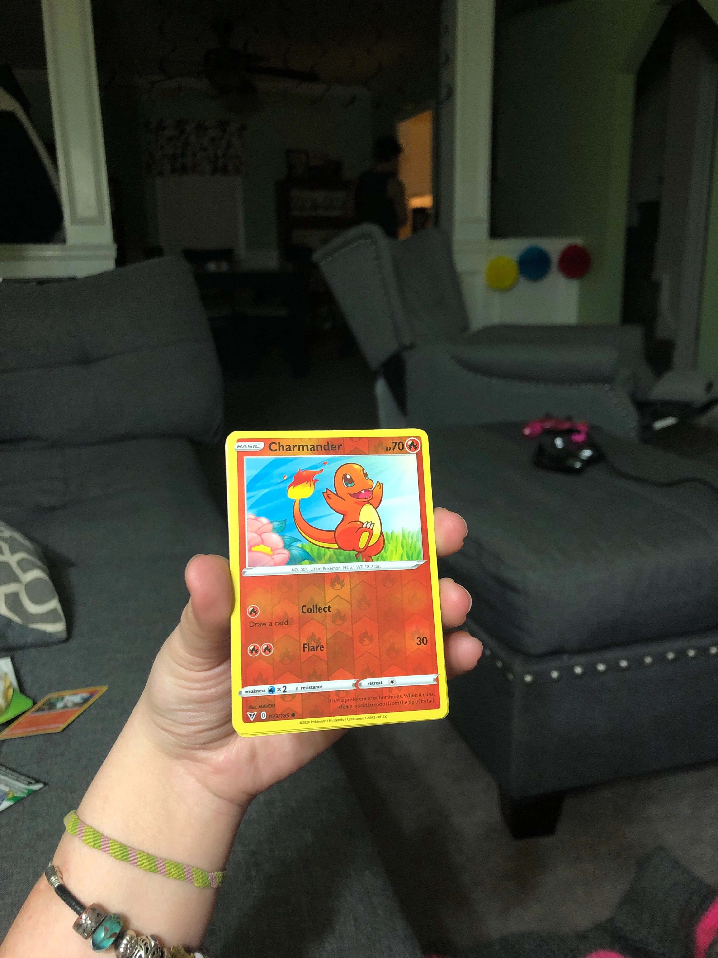 Pokemon Card 桌布 壁紙 — 一隻手在一個有灰色家具的舒適客廳前面拿著一張黃色的Charmander Pokémon卡。