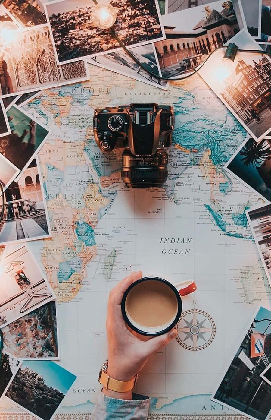 Travel Aesthetic Phone 桌布 壁紙 — 一隻手在一個被旅行照片和復古相機環繞的世界地圖上拿著一個咖啡杯。