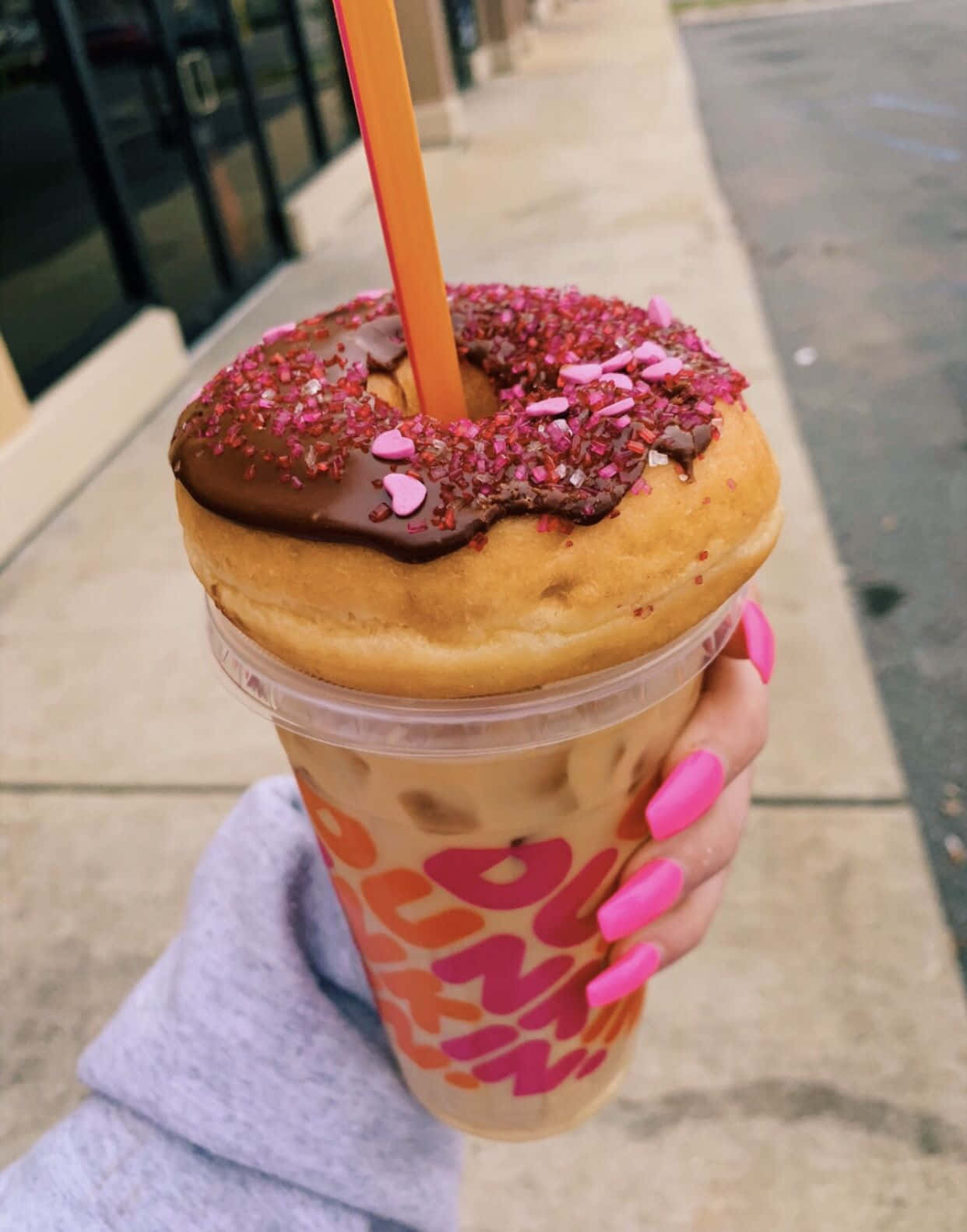 Dunkin Donuts 桌布 壁紙 — 一隻手在人行道上拿著一杯Dunkin'冰咖啡，上面有一個巧克力釉面甜甜圈和粉紅色糖粒。