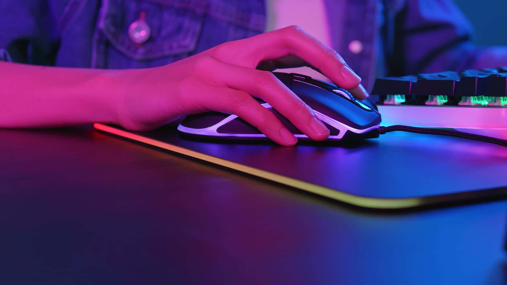 Gaming Mice 桌布 壁紙 — 一隻手在充滿活力的粉紅和藍色霓虹燈下，抓住一個發光鍵盤旁邊的遊戲滑鼠。