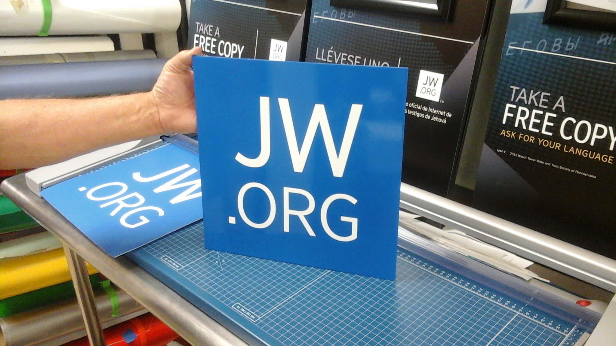sign 桌布 壁紙 — 一隻手在印刷店裡拿著一個藍色JW.ORG標誌，放在切割墊上，旁邊還有其他JW.ORG材料。