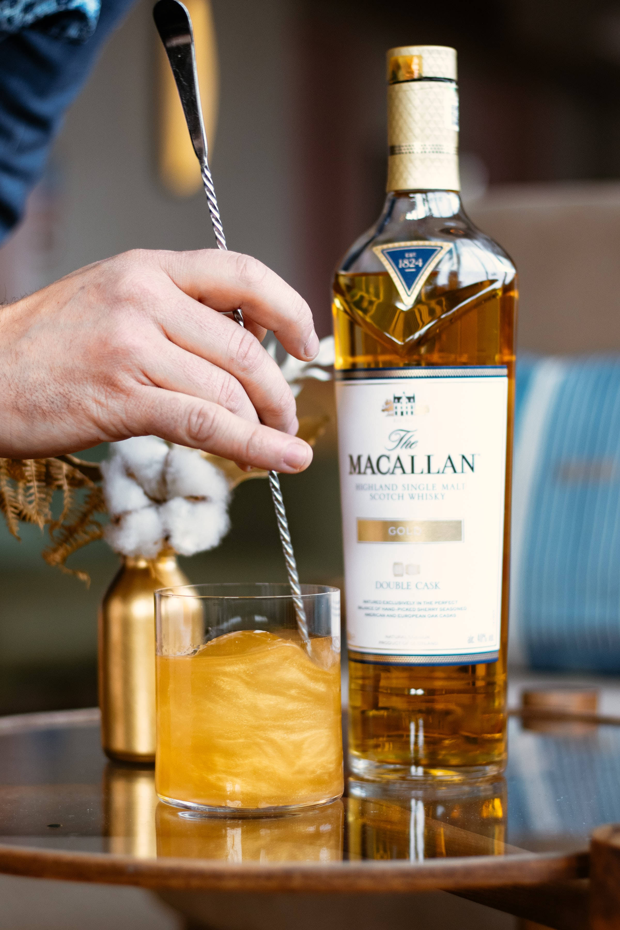alcohol 桌布 壁紙 — 一隻手在反光的木托盤上，於The Macallan Gold酒瓶旁邊攪拌一杯金色的威士忌雞尾酒。