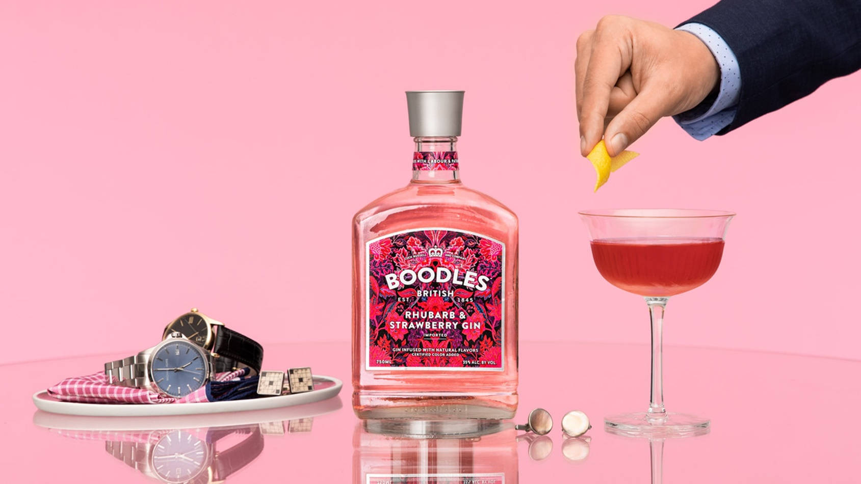 alcohol 桌布 壁紙 — 一隻手在反光的粉紅色表面上，於一瓶Boodles Rhubarb & Strawberry Gin和奢華手錶旁邊裝飾一杯粉紅色琴酒雞尾酒。