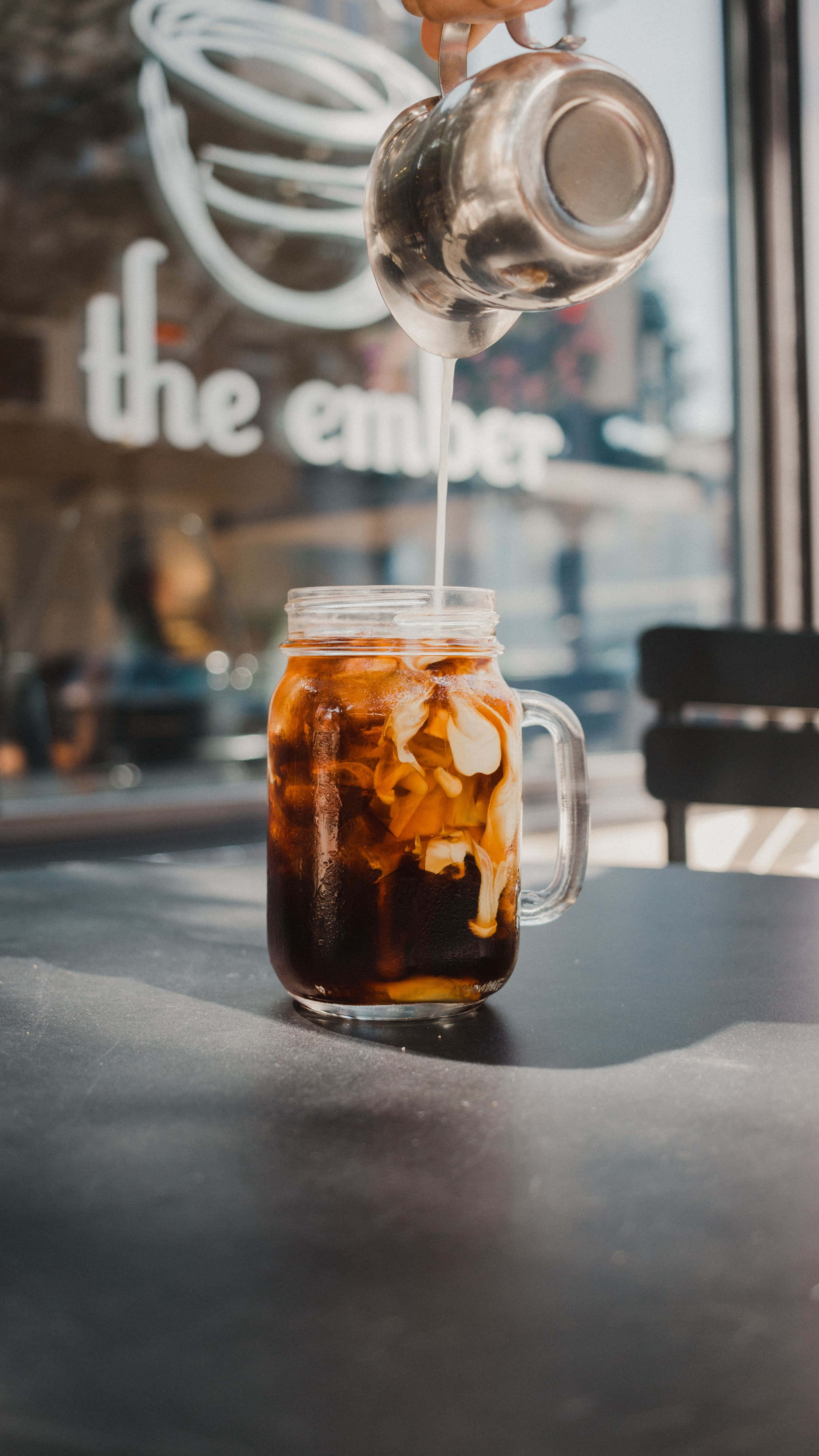 Iced Coffee 桌布 壁紙 — 一隻手在咖啡店將奶油牛奶倒入一杯冰咖啡中，背景是模糊的商店招牌。