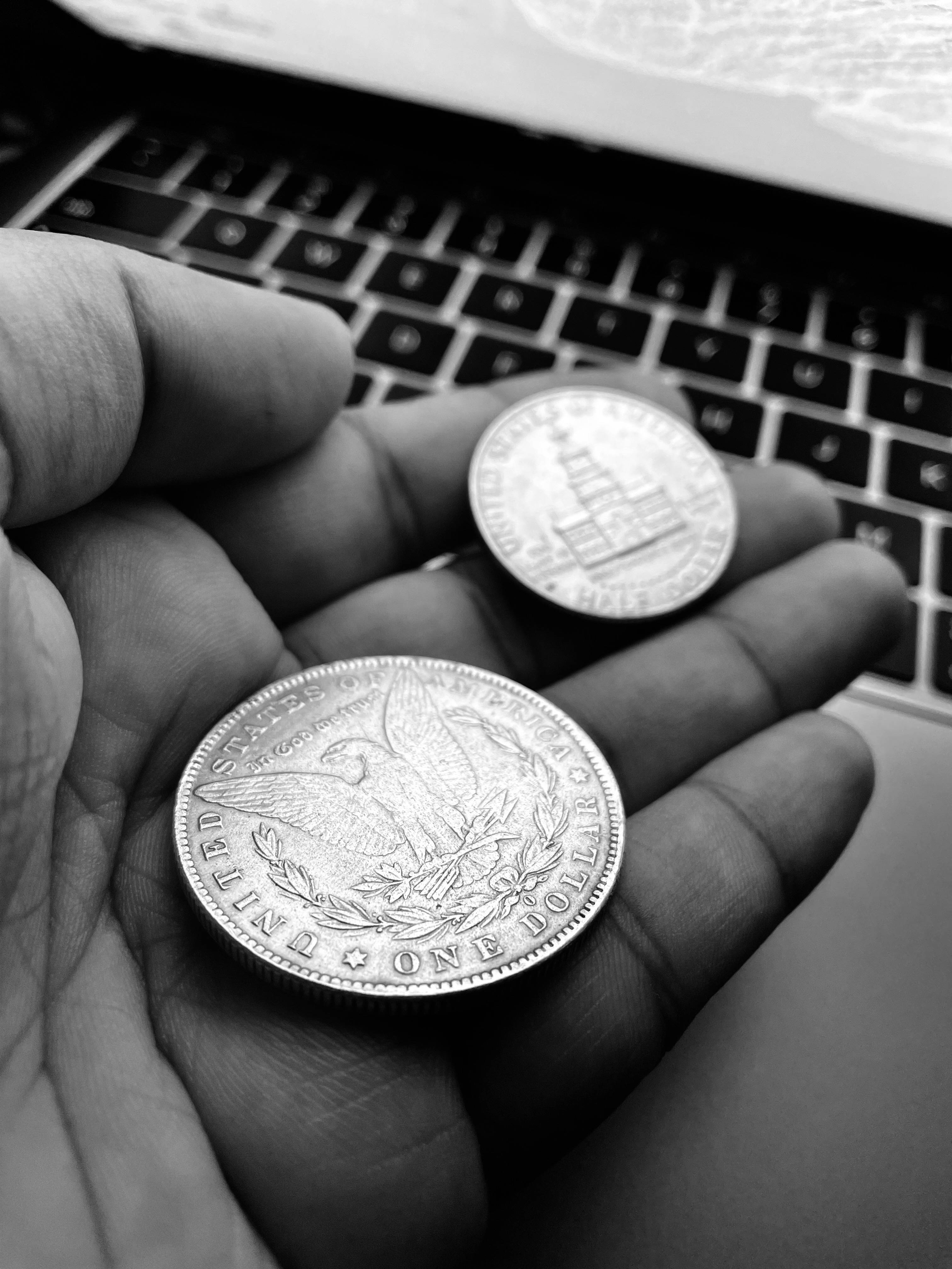 金錢 iPhone 桌布 壁紙 — 一隻手在單色特寫中，於筆記型電腦鍵盤前拿著兩枚精細的銀幣。