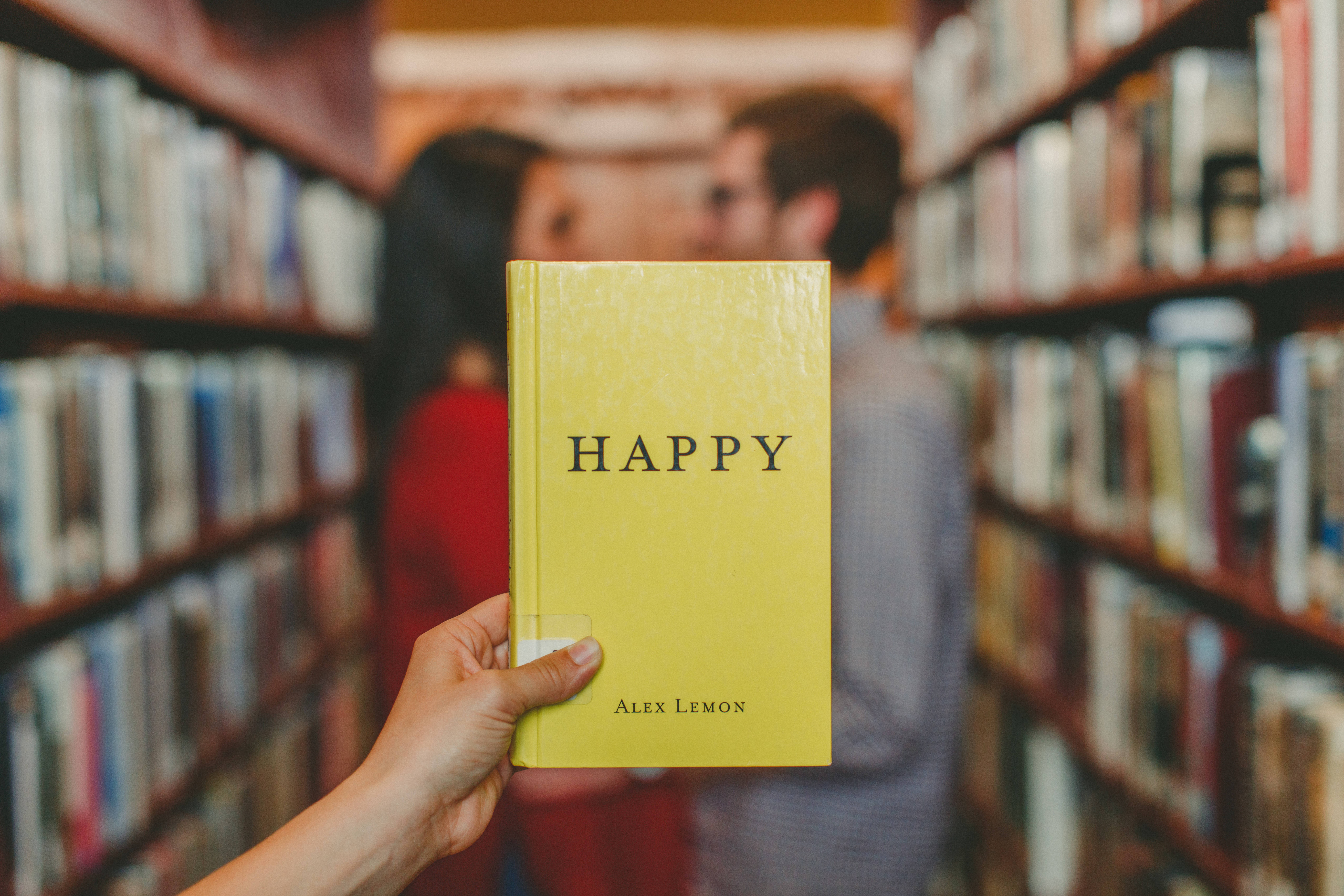 快樂 桌布 壁紙 — 一隻手在圖書館走道中拿著一本名為「HAPPY」的黃色書，背景模糊中有一對情侶在接吻。