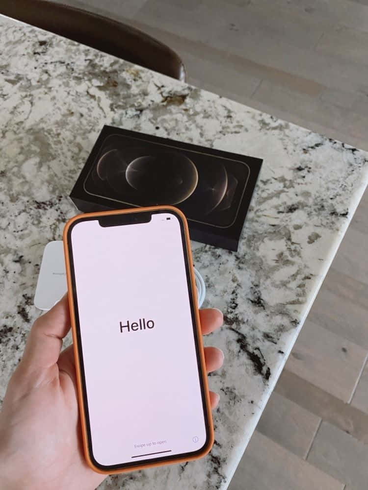 iPhone 12 桌布 壁紙 — 一隻手在大理石檯面上，用橙色機殼的新iPhone在原裝盒上方顯示Hello畫面。