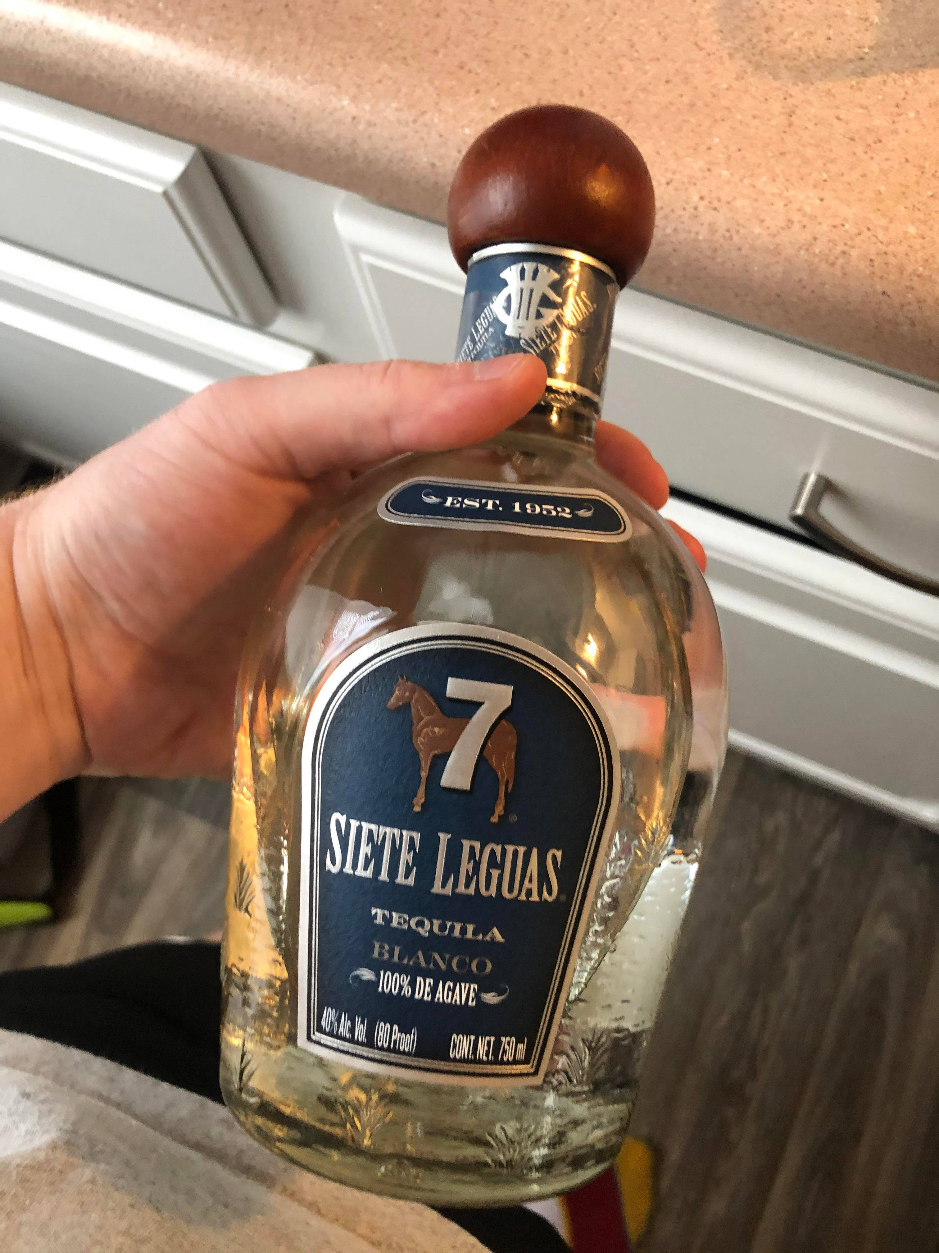 雙螢幕桌布 桌布 壁紙 — 一隻手在廚房檯面上拿著一瓶帶有木塞的透明 Siete Leguas Blanco 龍舌蘭酒。