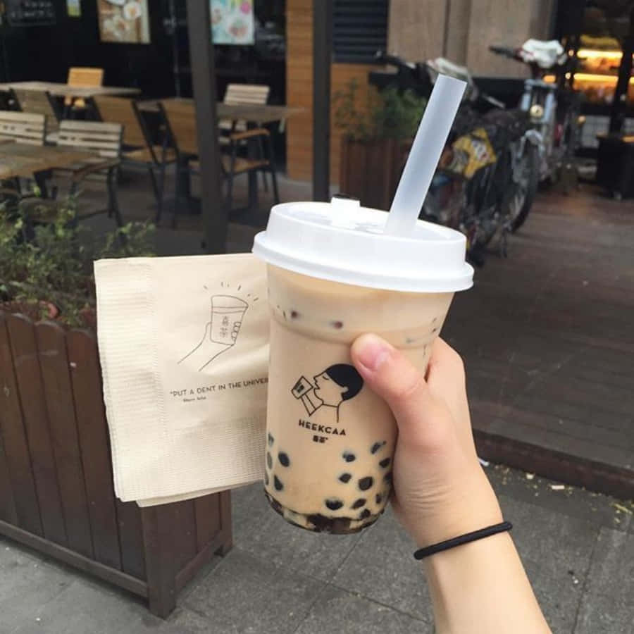 Boba Tea 桌布 壁紙 — 一隻手在戶外咖啡廳座位區附近，拿著一杯奶油味珍珠奶茶（有黑色木薯珍珠）和一張有品牌的紙巾。