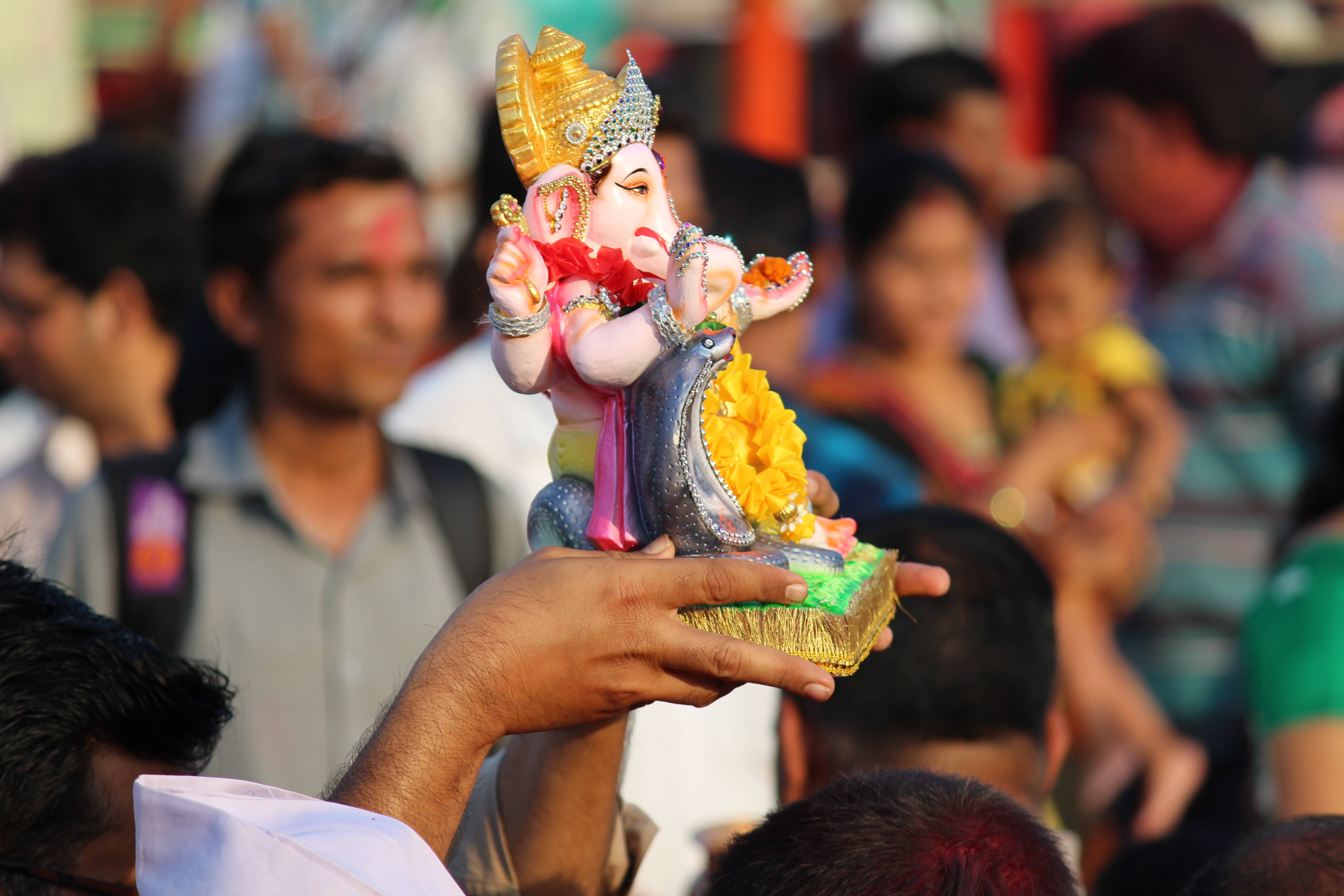 ganesh 4K 桌布 壁紙 — 一隻手在擁擠的節日中拿著一個色彩繽紛、華麗的象神雕像，飾有黃金和鮮花。