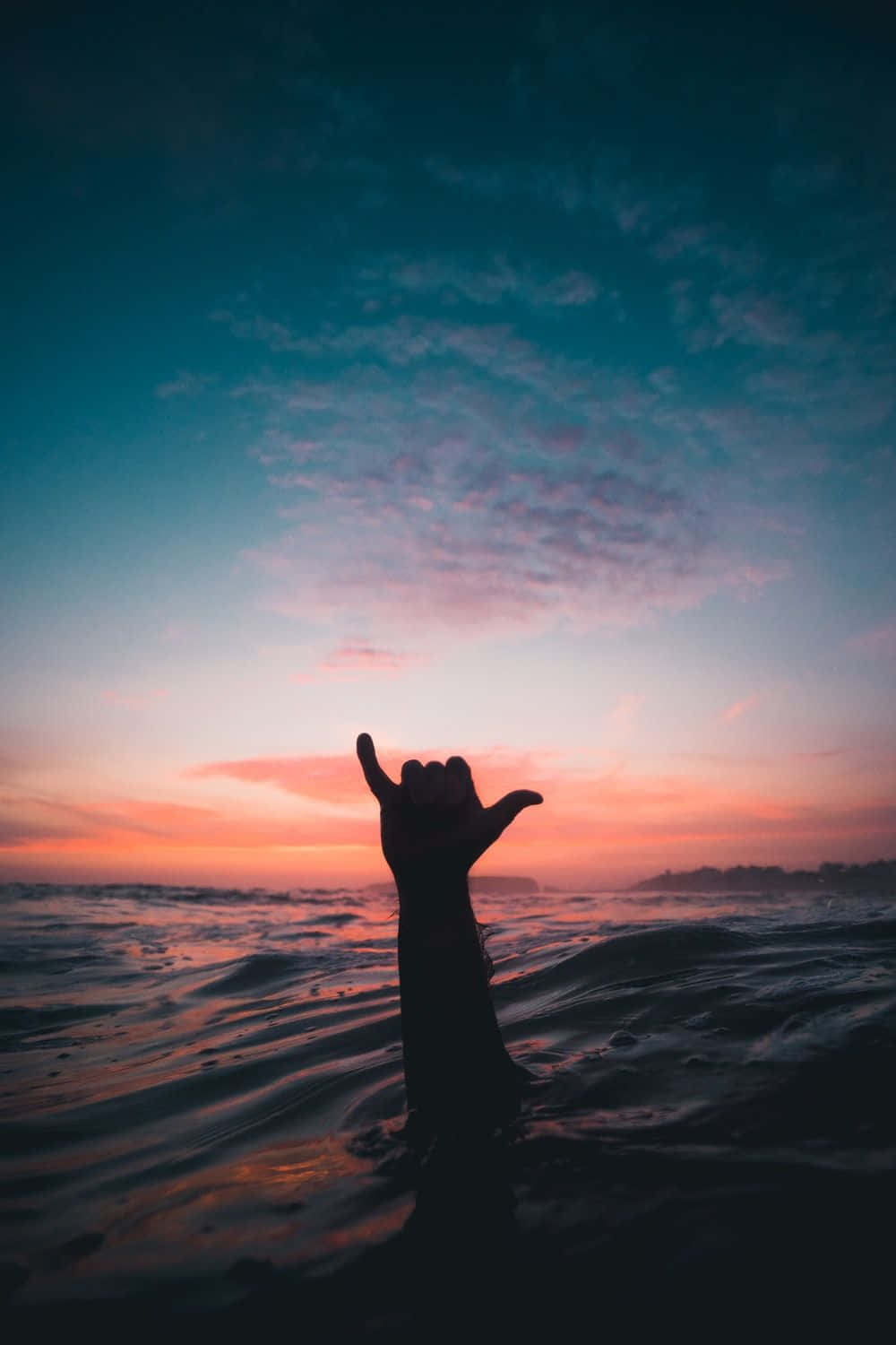 Surfing iPhone 桌布 壁紙 — 一隻手在日落時從海浪中伸出，對著充滿活力的粉紅色和藍綠色天空做出夏卡手勢。